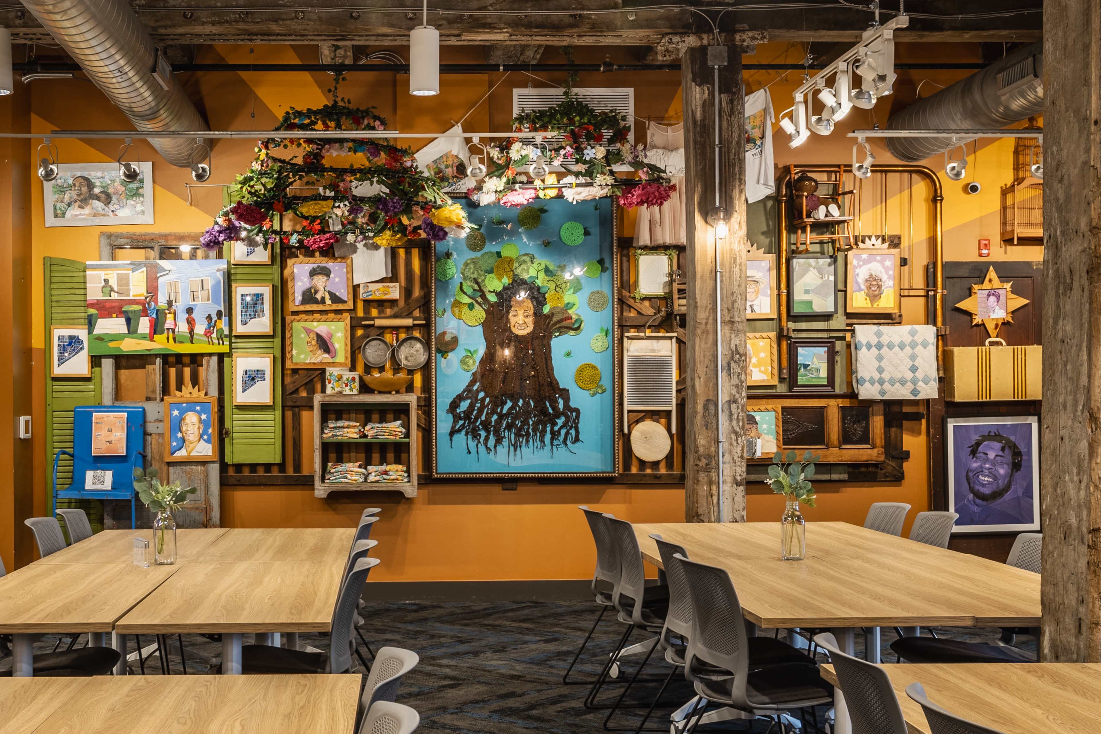 The image depicts a vibrant community space with wooden beams, colorful artwork on the walls, and several tables set up for gatherings.