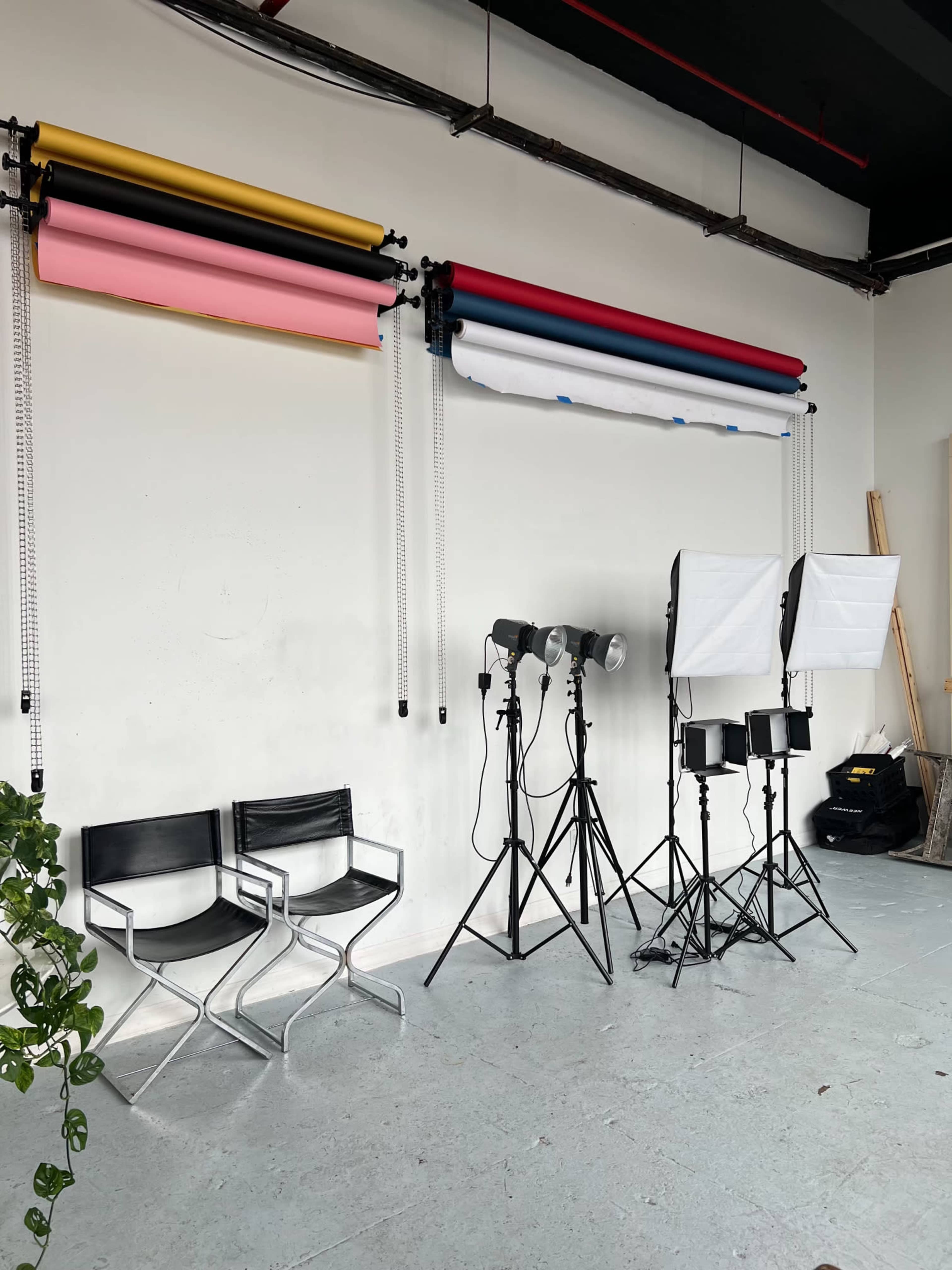 A photography studio setup featuring rolled backdrops in various colors, two black chairs, and multiple lighting stands arranged beside a wall.
