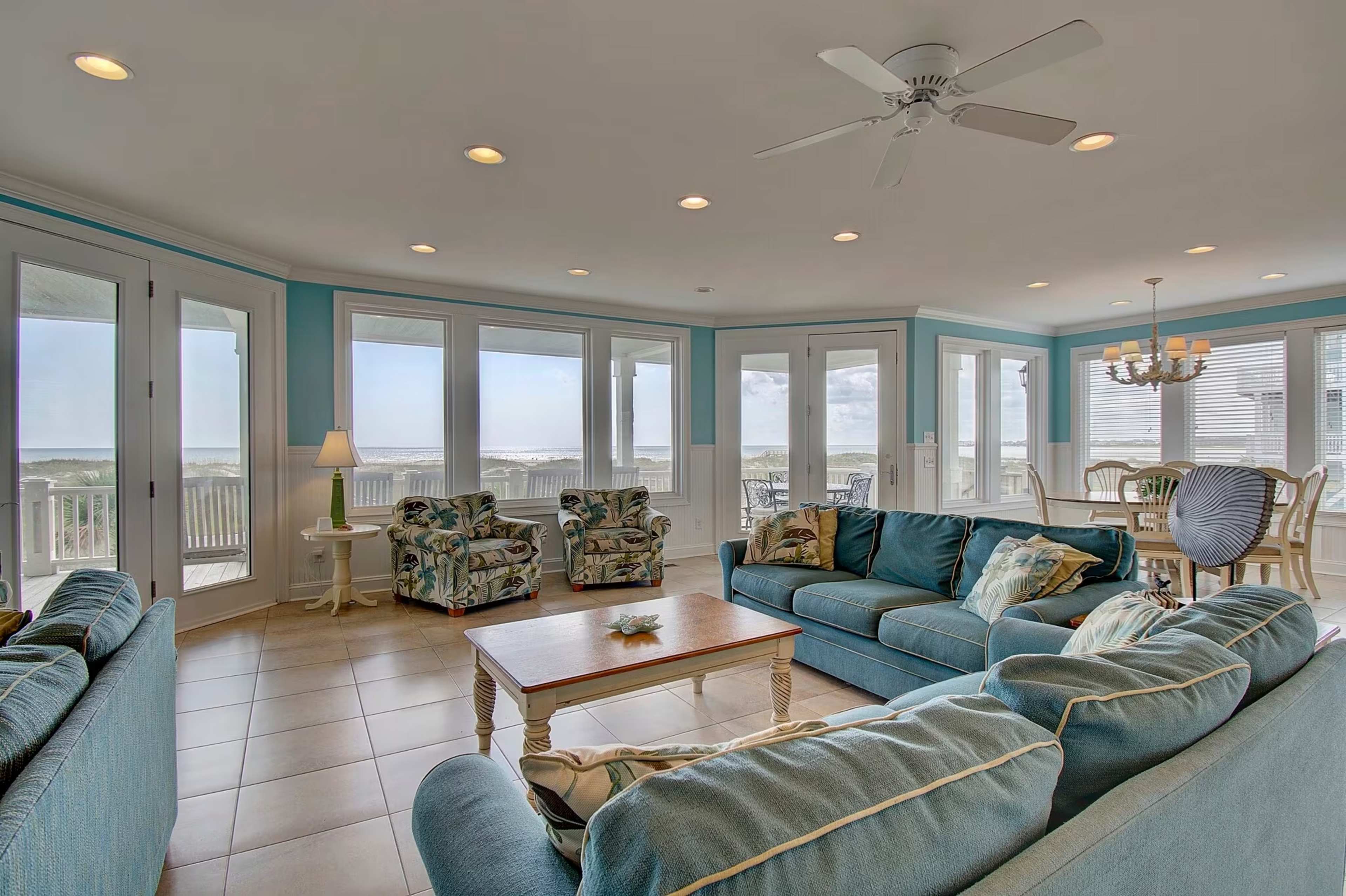 The living room features a spacious arrangement of blue couches and chairs, with large windows offering a view of the ocean.