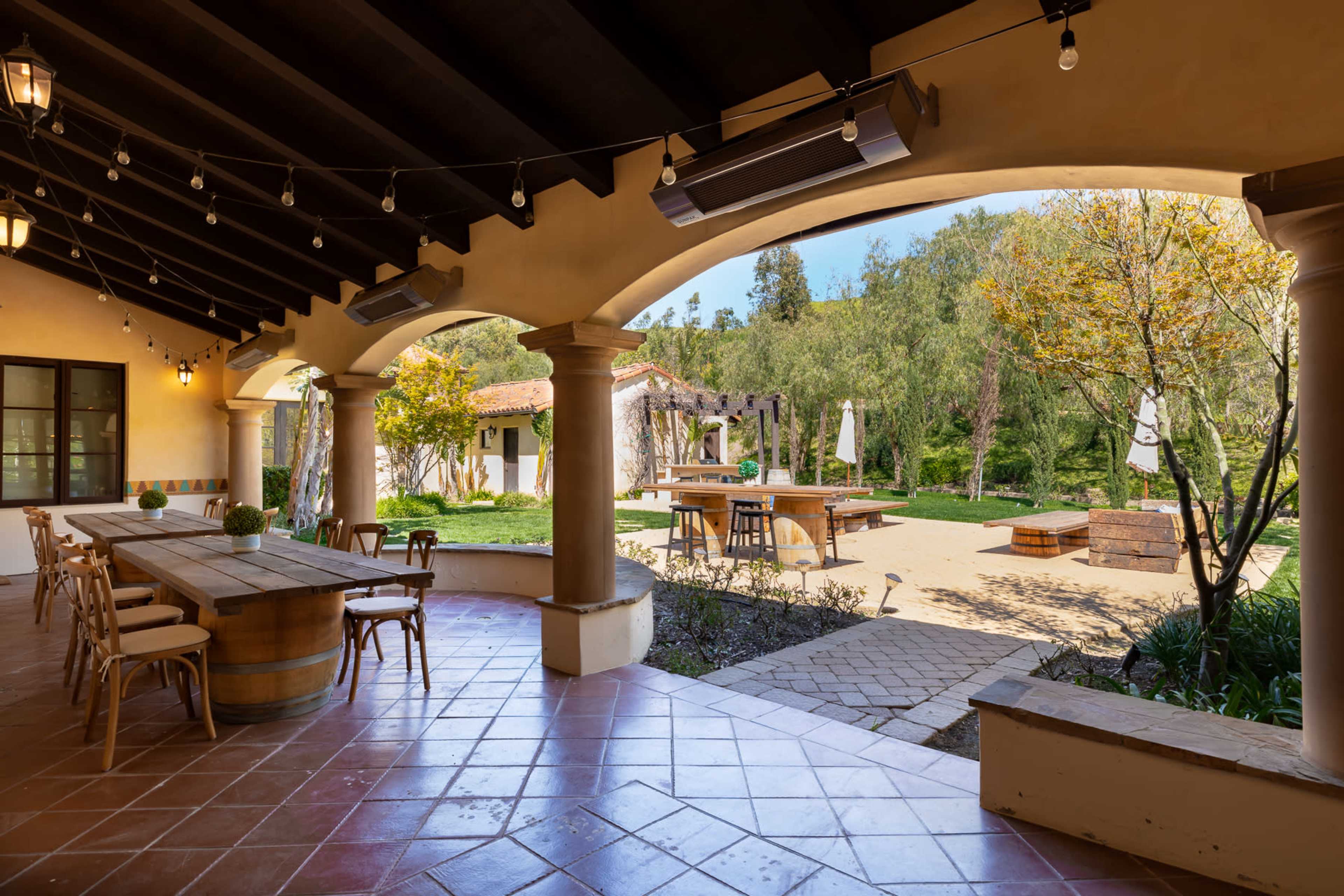 An outdoor patio area featuring a long wooden table, chairs, and lush greenery in the background.