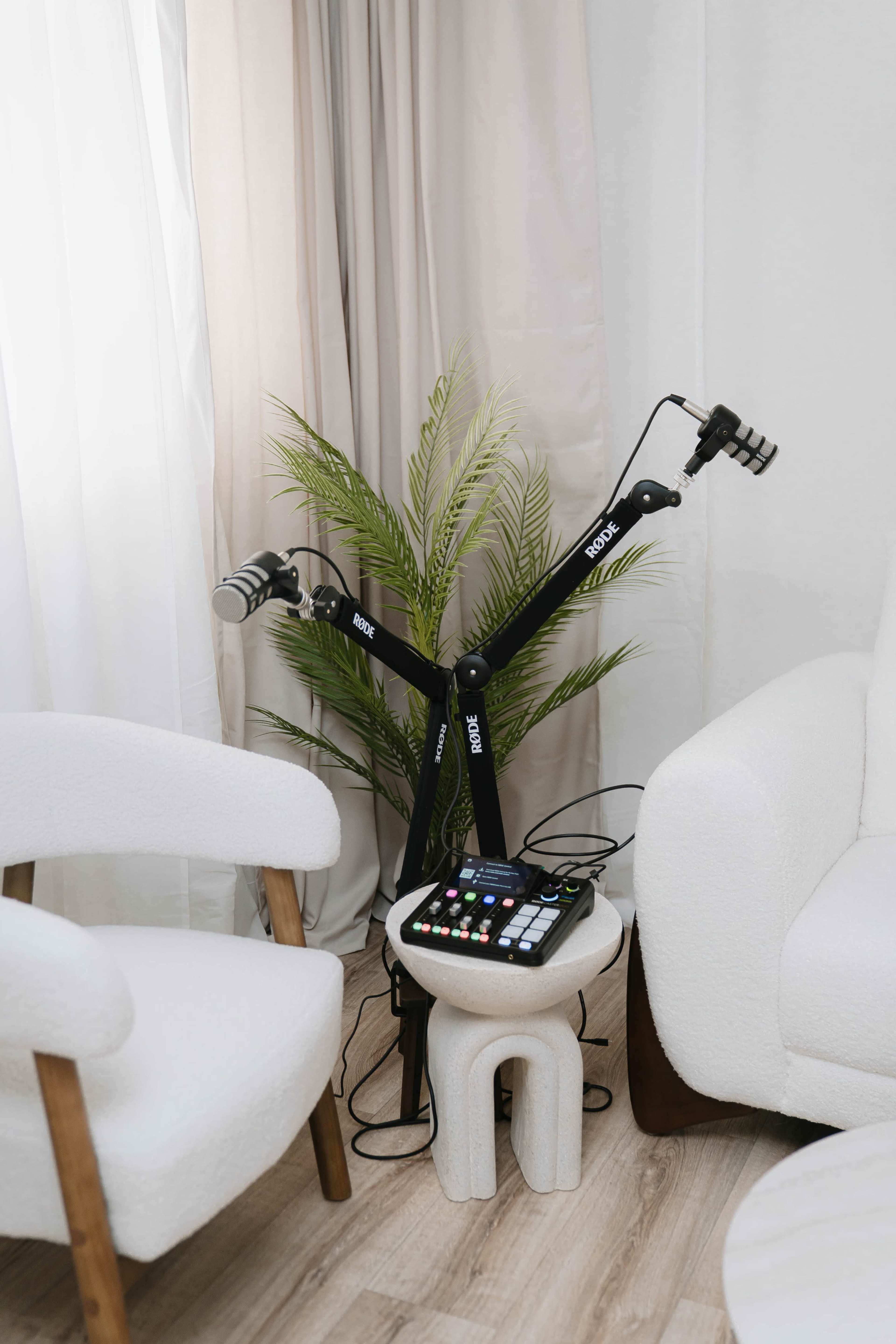 A microphone stand is positioned next to a small, round stool with a multi-functional audio device on top, beside two white chairs in a bright room with light curtains and a potted plant.