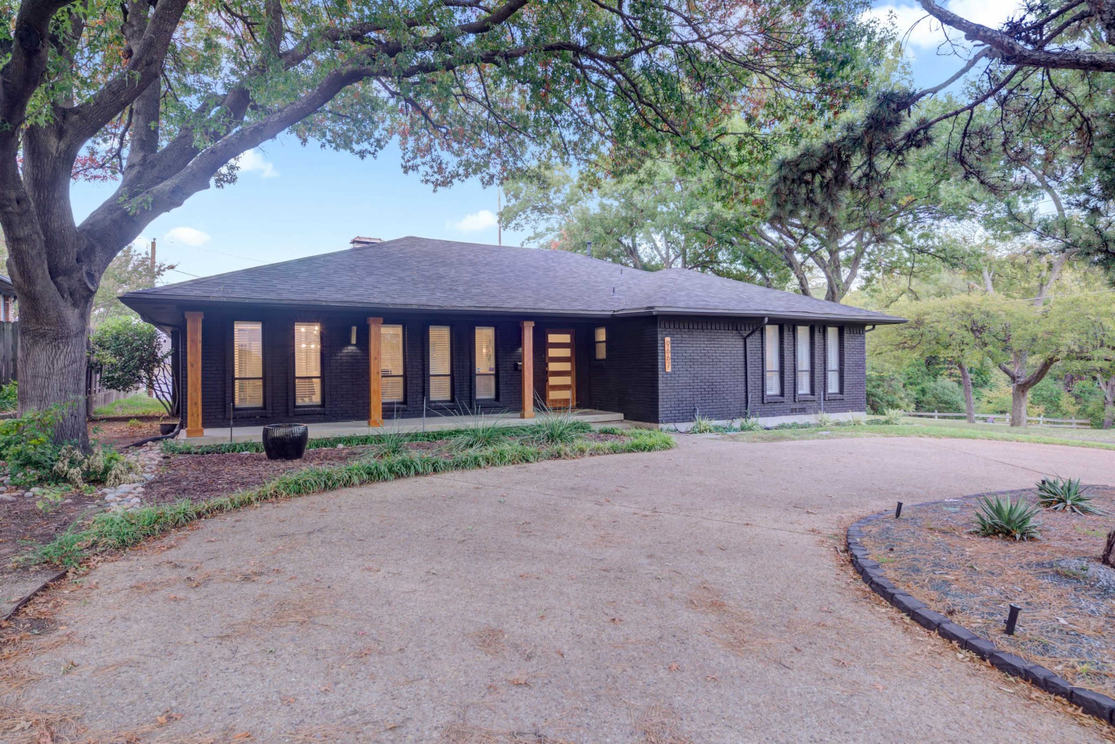 The image shows a modern, single-story black brick house with a gravel driveway surrounded by trees and landscaping.