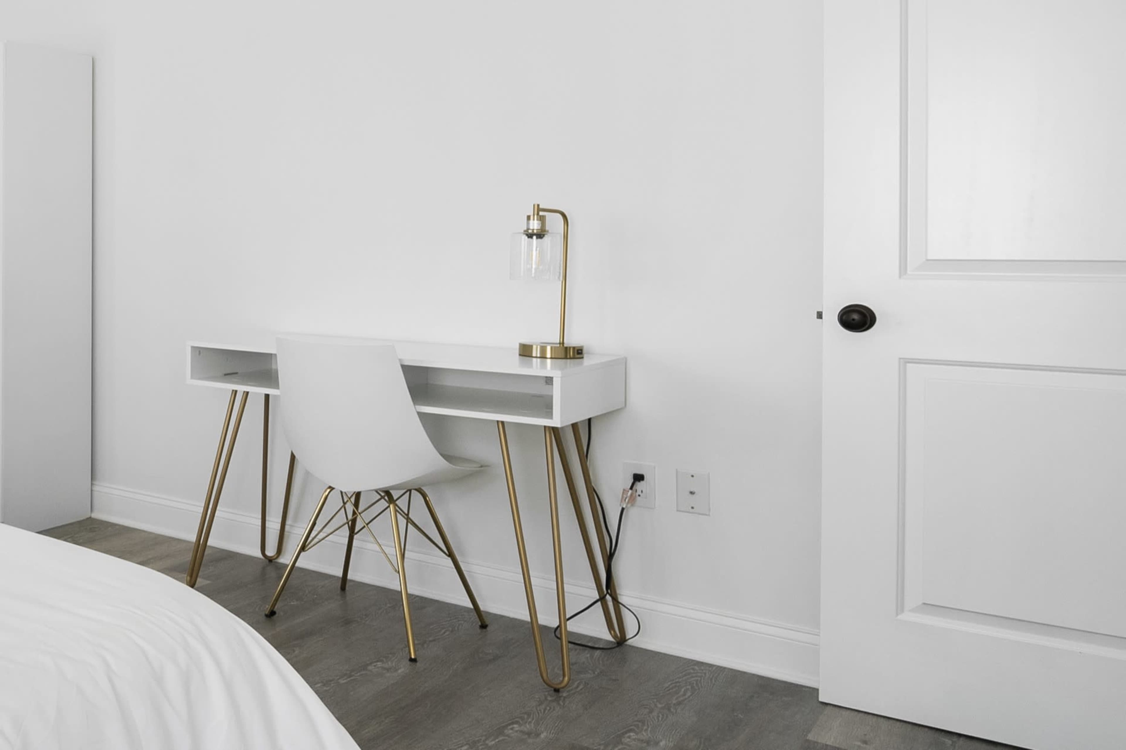The image shows a minimalist workspace featuring a white desk with gold legs and a matching lamp beside a closed white door.