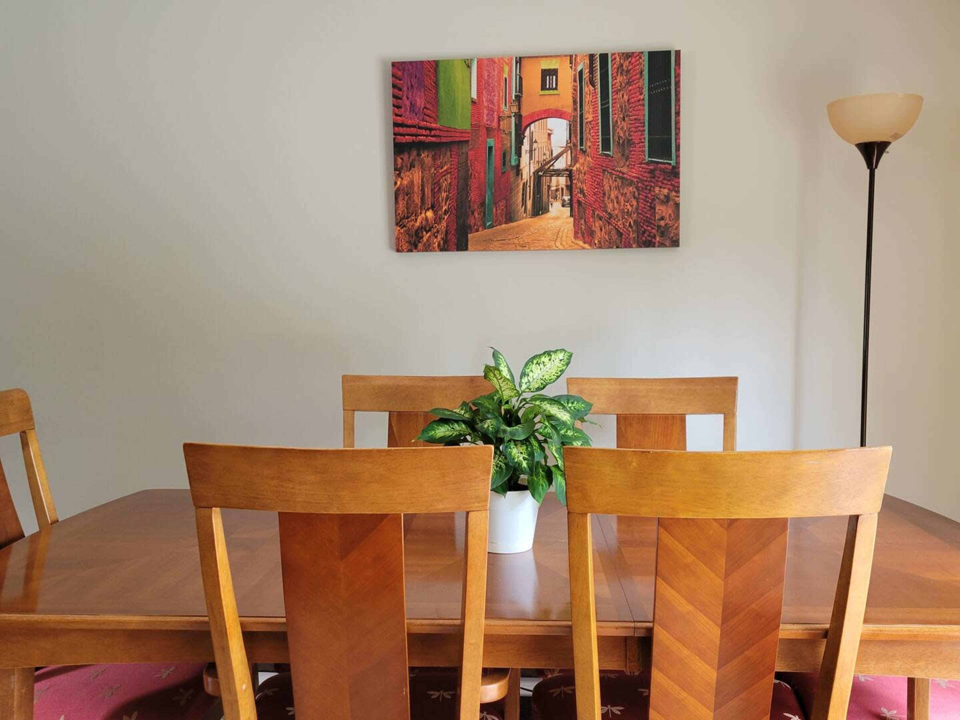A wooden dining table with chairs is set in a room featuring a wall-hanging photo of a narrow street, with a lamp in the corner.