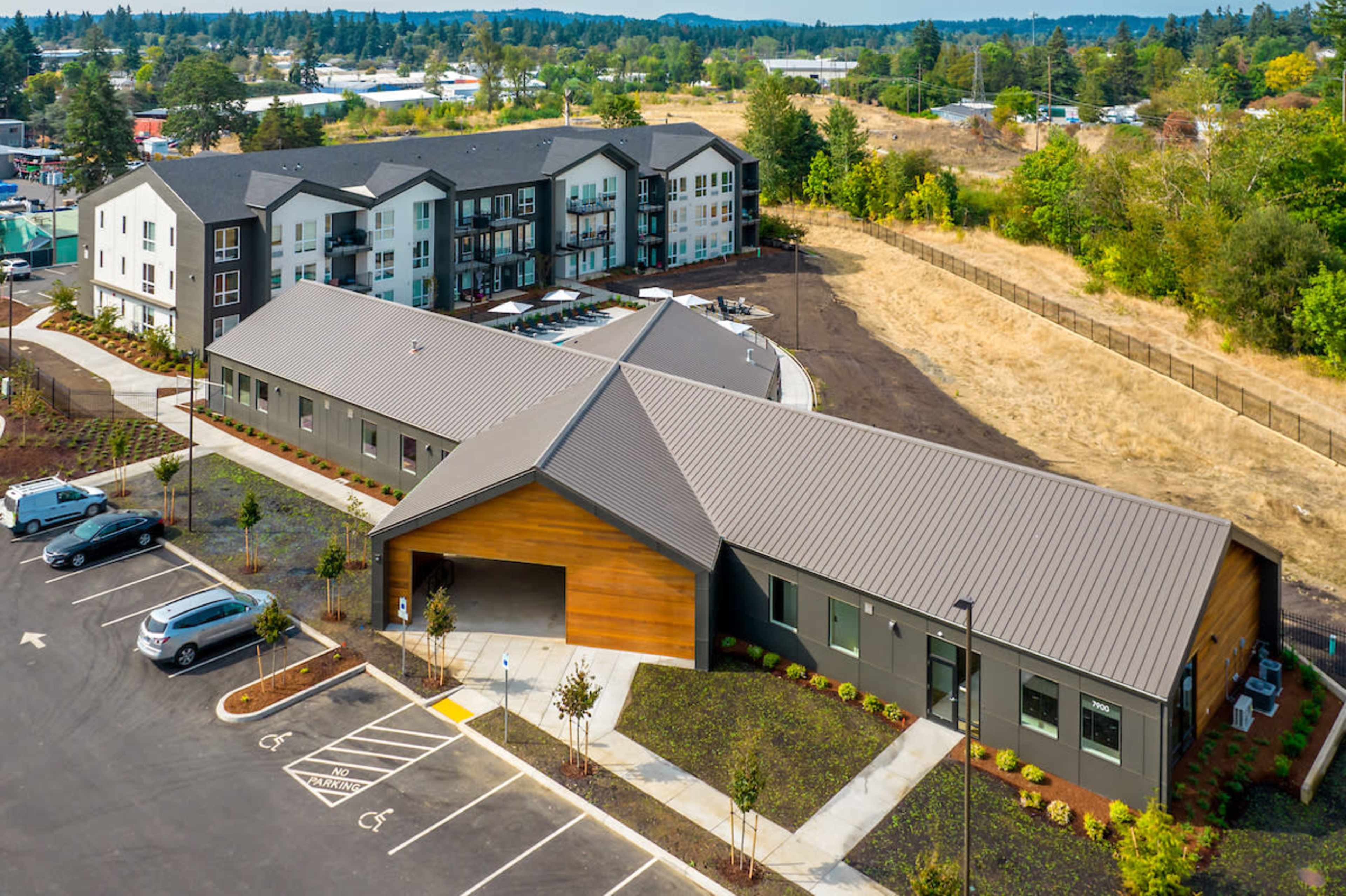 The image shows a modern multi-story apartment complex situated next to a single-story building, surrounded by paved parking and landscaped grounds.