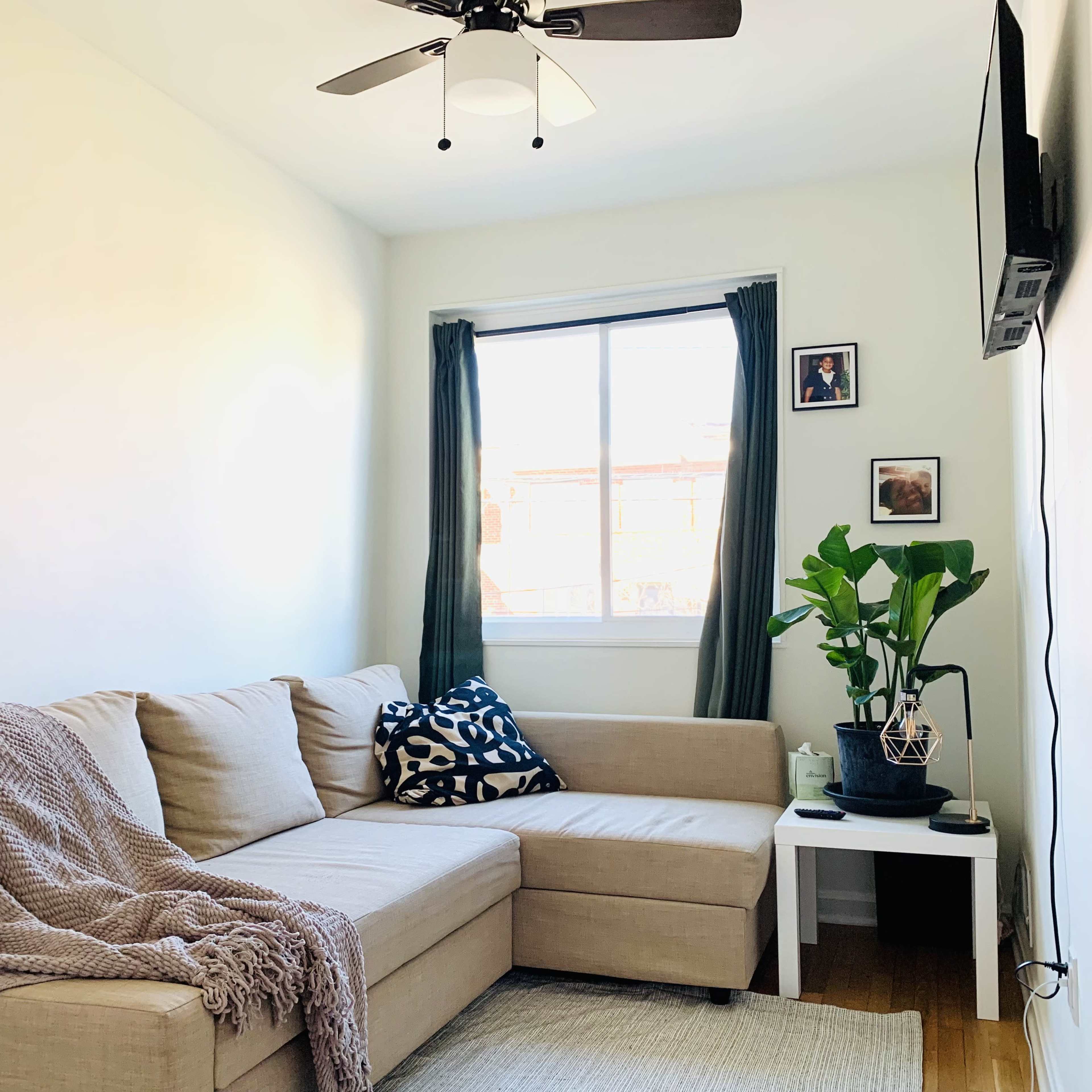 A light, airy living room features a beige sectional sofa, a potted plant, and framed photographs on the wall.