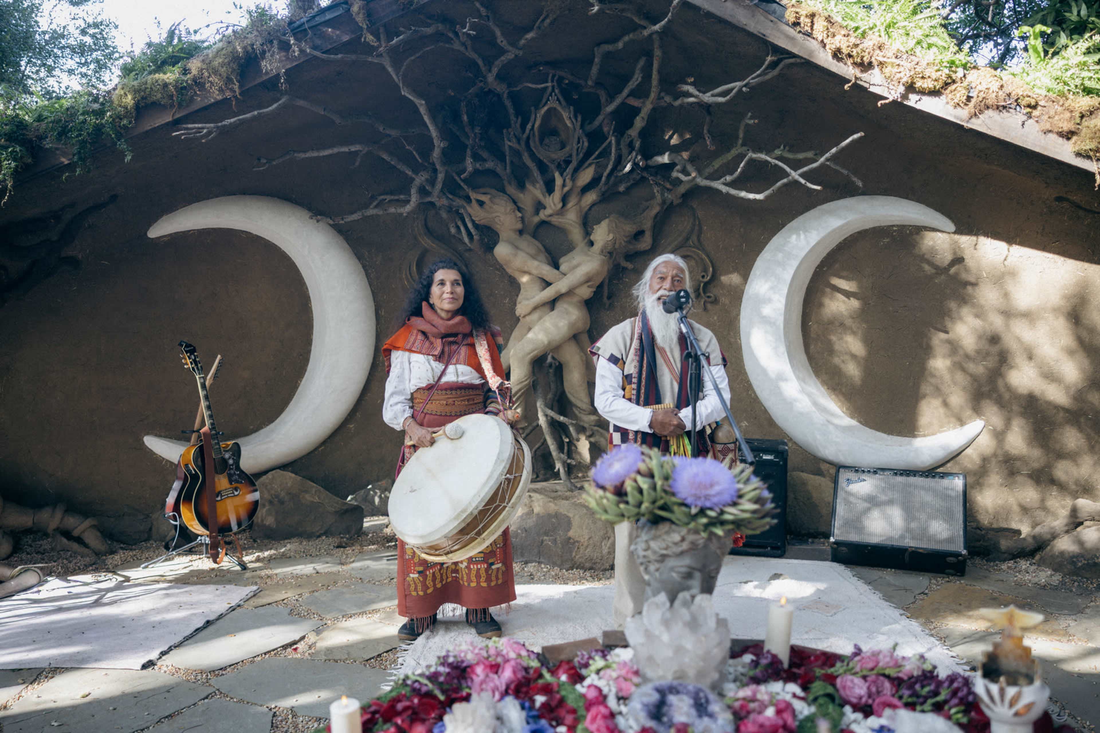 Two musicians perform in front of a large, decorative wall featuring crescent moons, with one playing a drum and the other singing into a microphone, surrounded by floral arrangements and instruments.