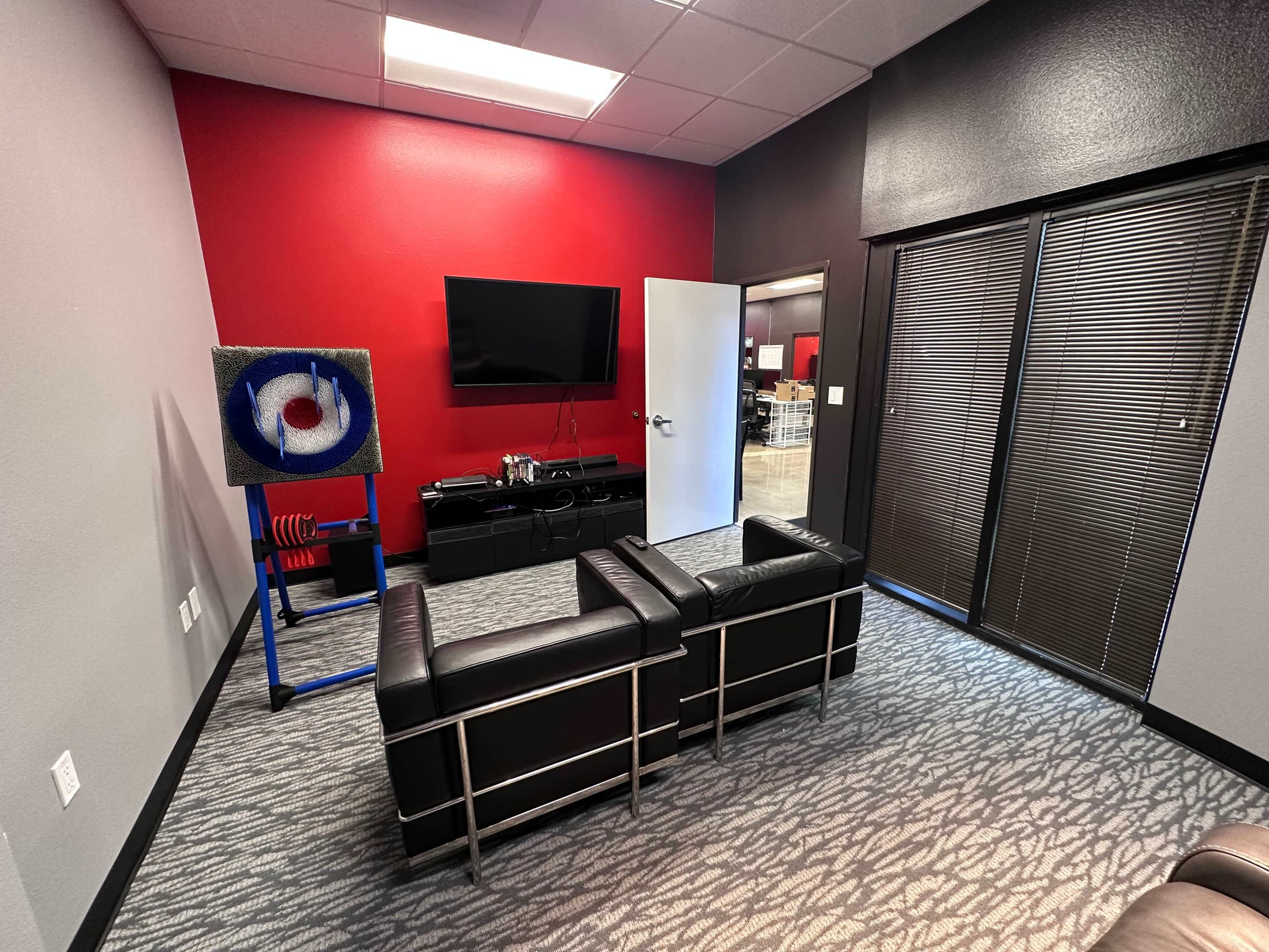 A modern office lounge with two black chairs facing a television and a colorful target on an easel against a red accent wall.