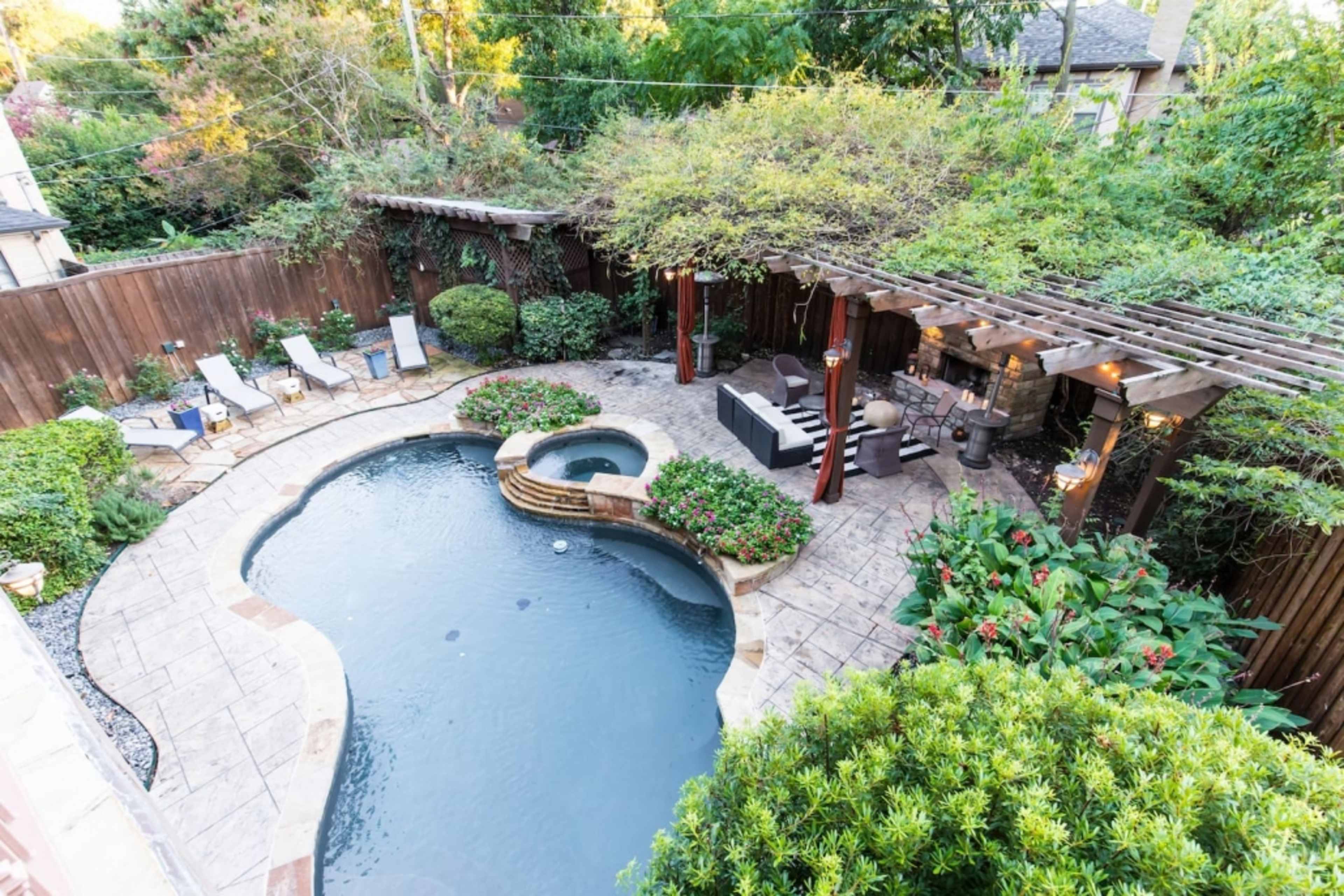 A well-landscaped backyard with a curved swimming pool, a spa area, and outdoor seating beneath a pergola surrounded by greenery.