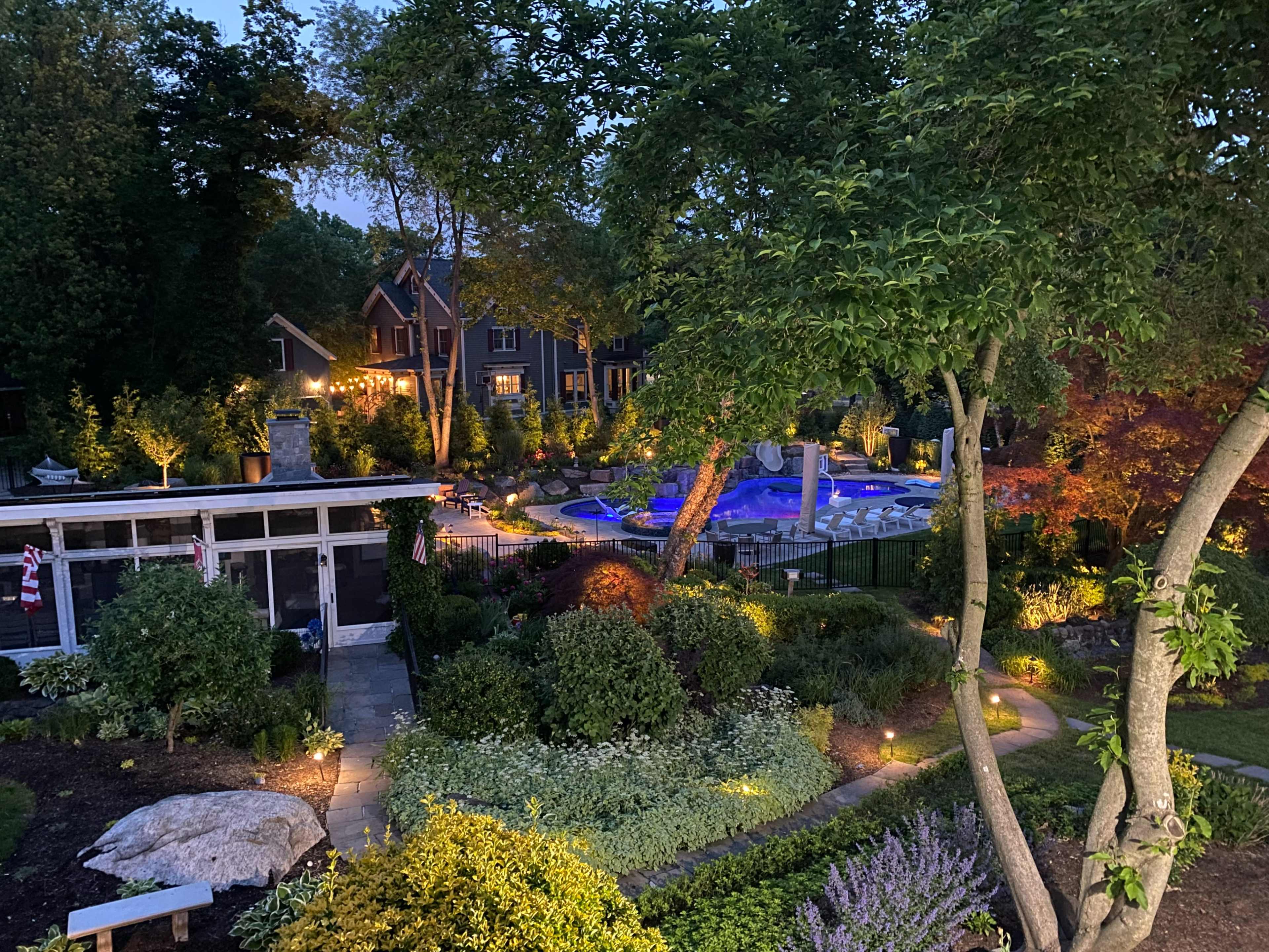 A landscaped garden with a swimming pool is illuminated at night, surrounded by trees and residential homes.