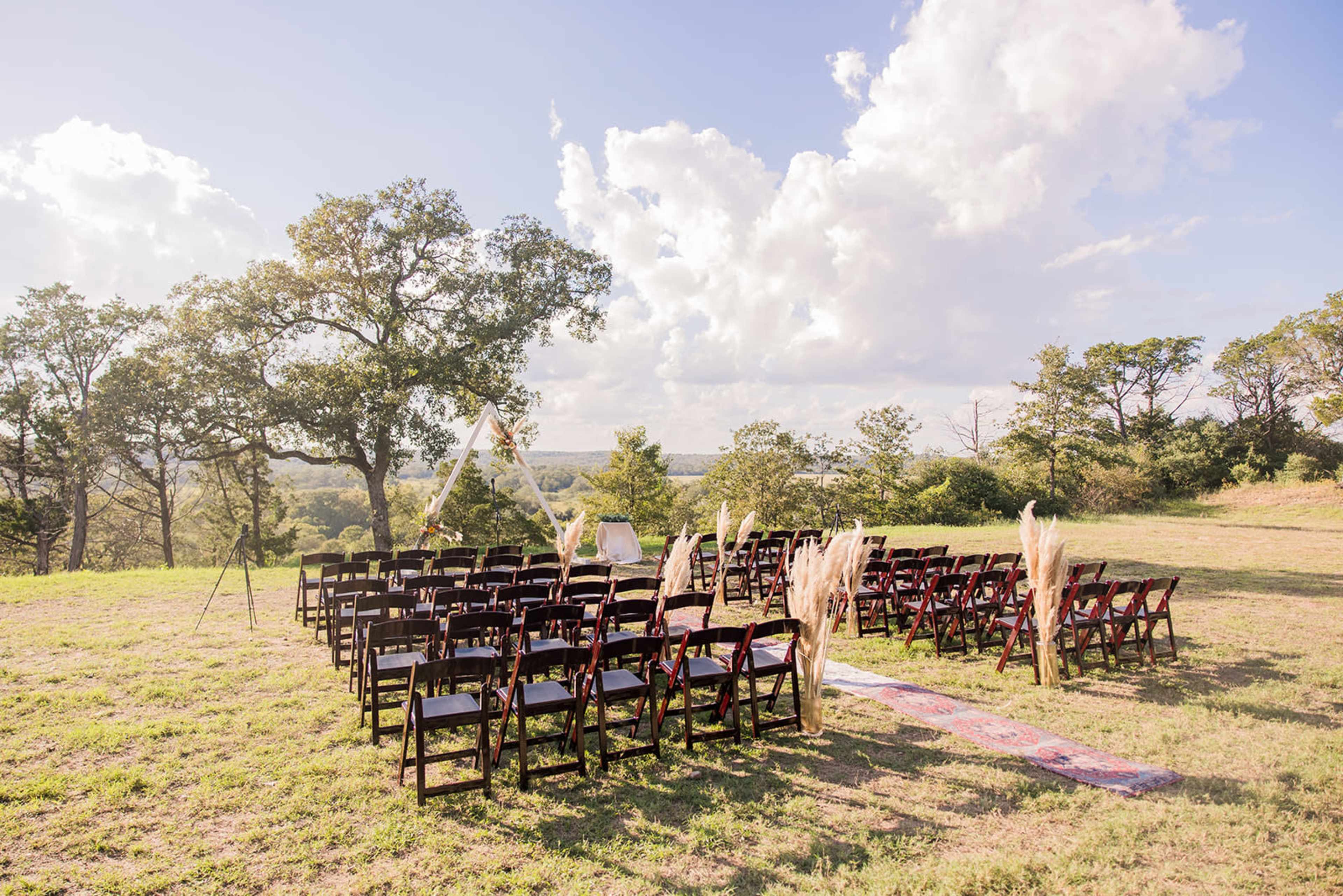A wedding ceremony setup with rows of dark wooden chairs arranged on a grassy hillside under a partly cloudy sky.