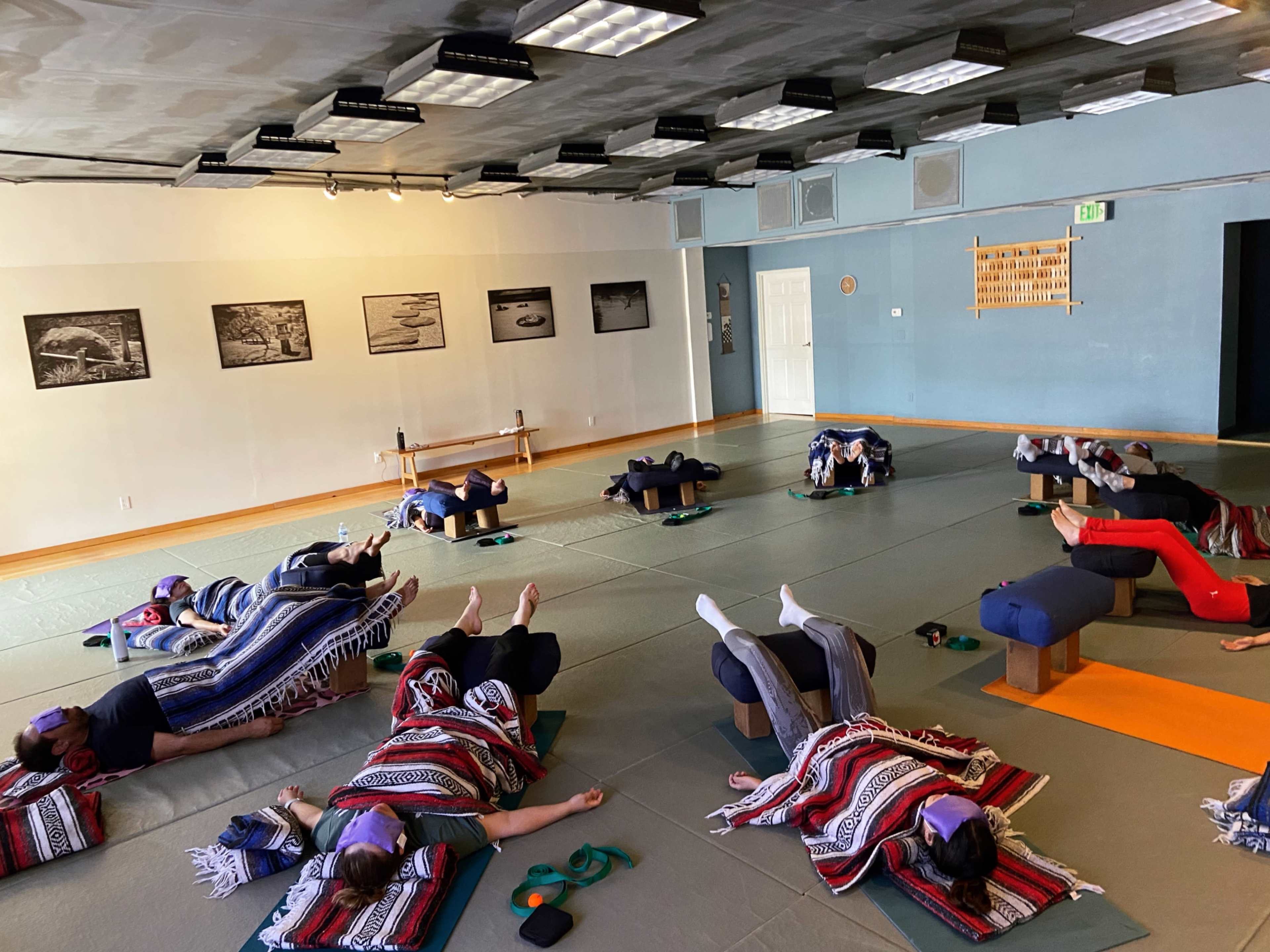 A group of individuals practices relaxation exercises on mats in a yoga studio, some covered with blankets and using bolsters for support.
