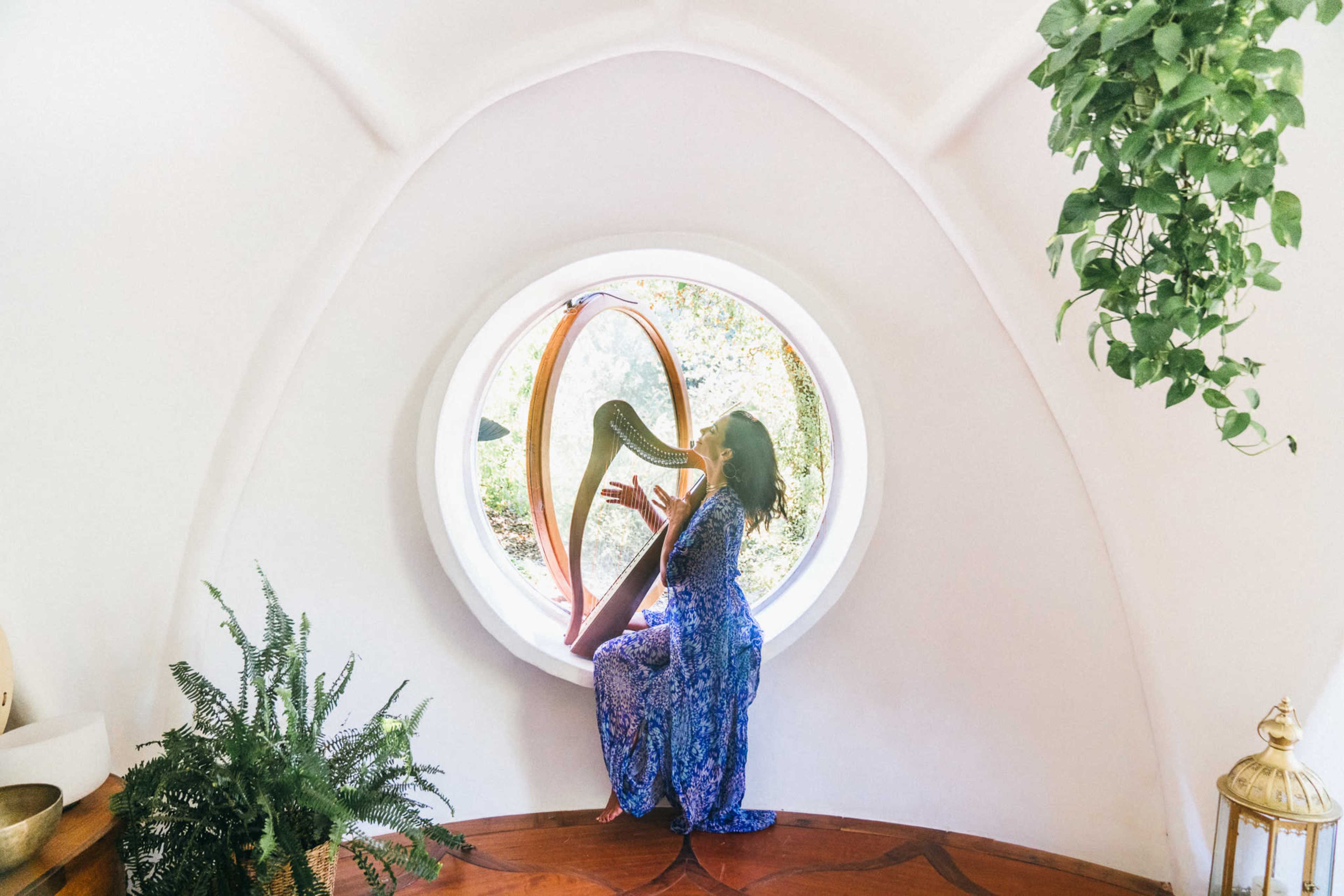 A woman in a blue patterned dress plays a harp while seated in a circular window surrounded by greenery.
