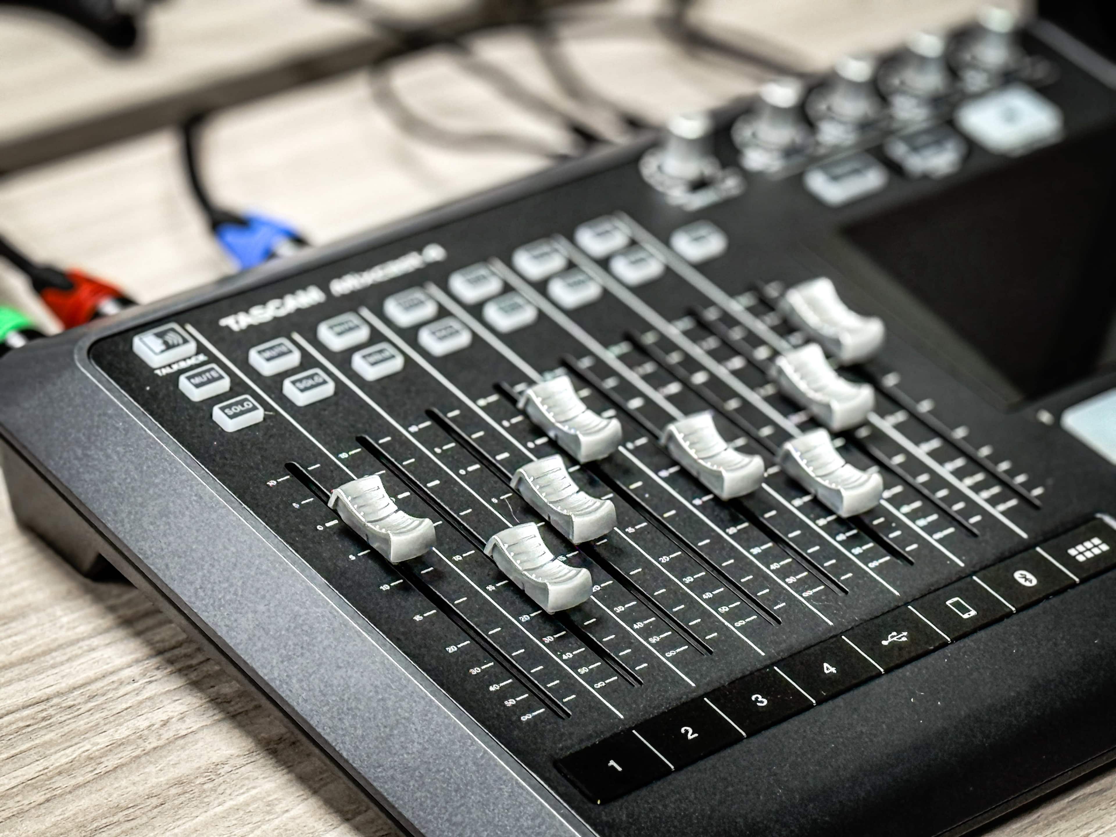 The image shows a TASCAM audio mixer with multiple sliding faders and various buttons on its surface.