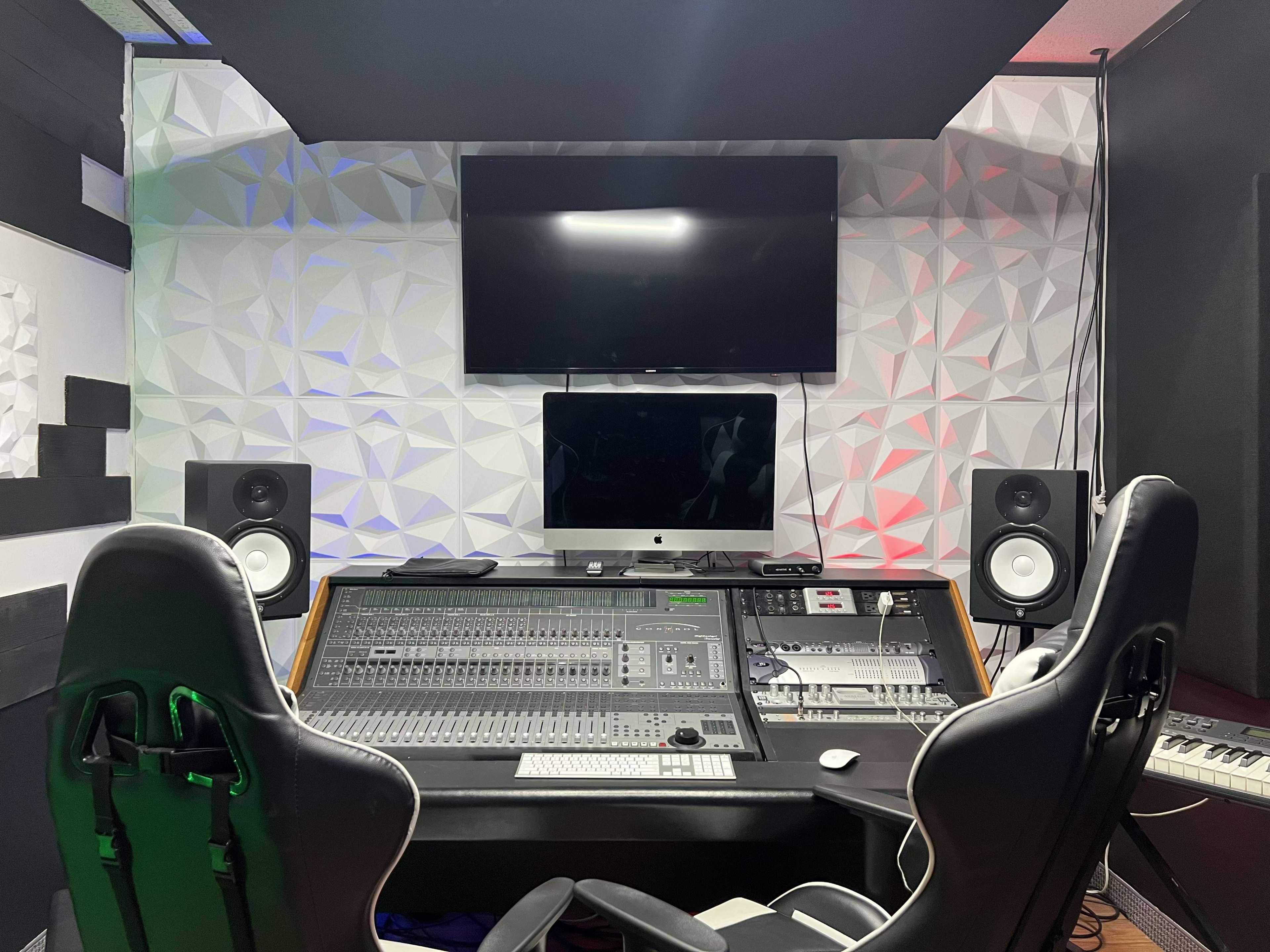 A music recording studio featuring a large mixing console, two monitors, and a television mounted on the wall.