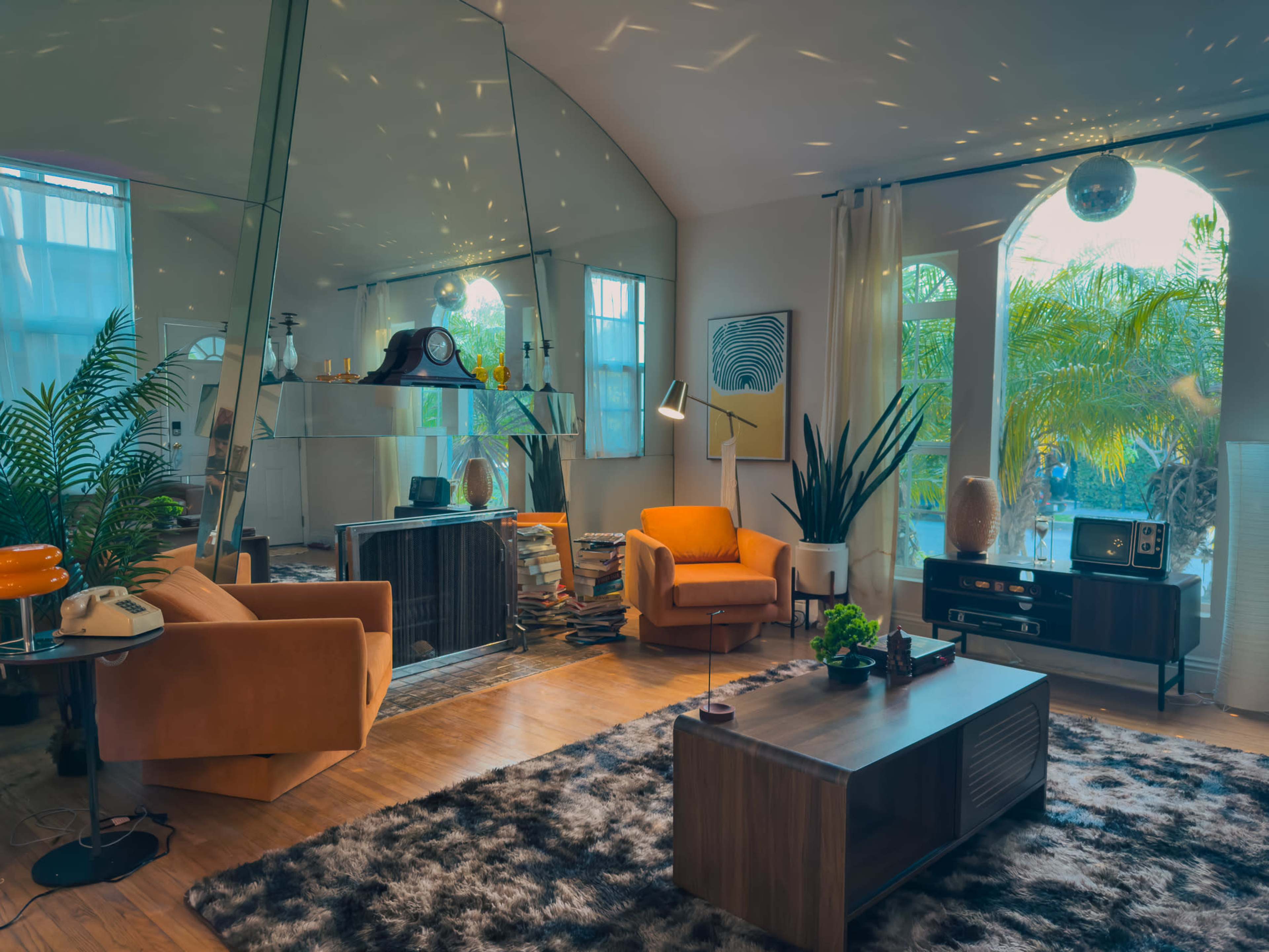 The image shows a stylish living room featuring orange armchairs, a mirrored wall, a large area rug, and a variety of plants.