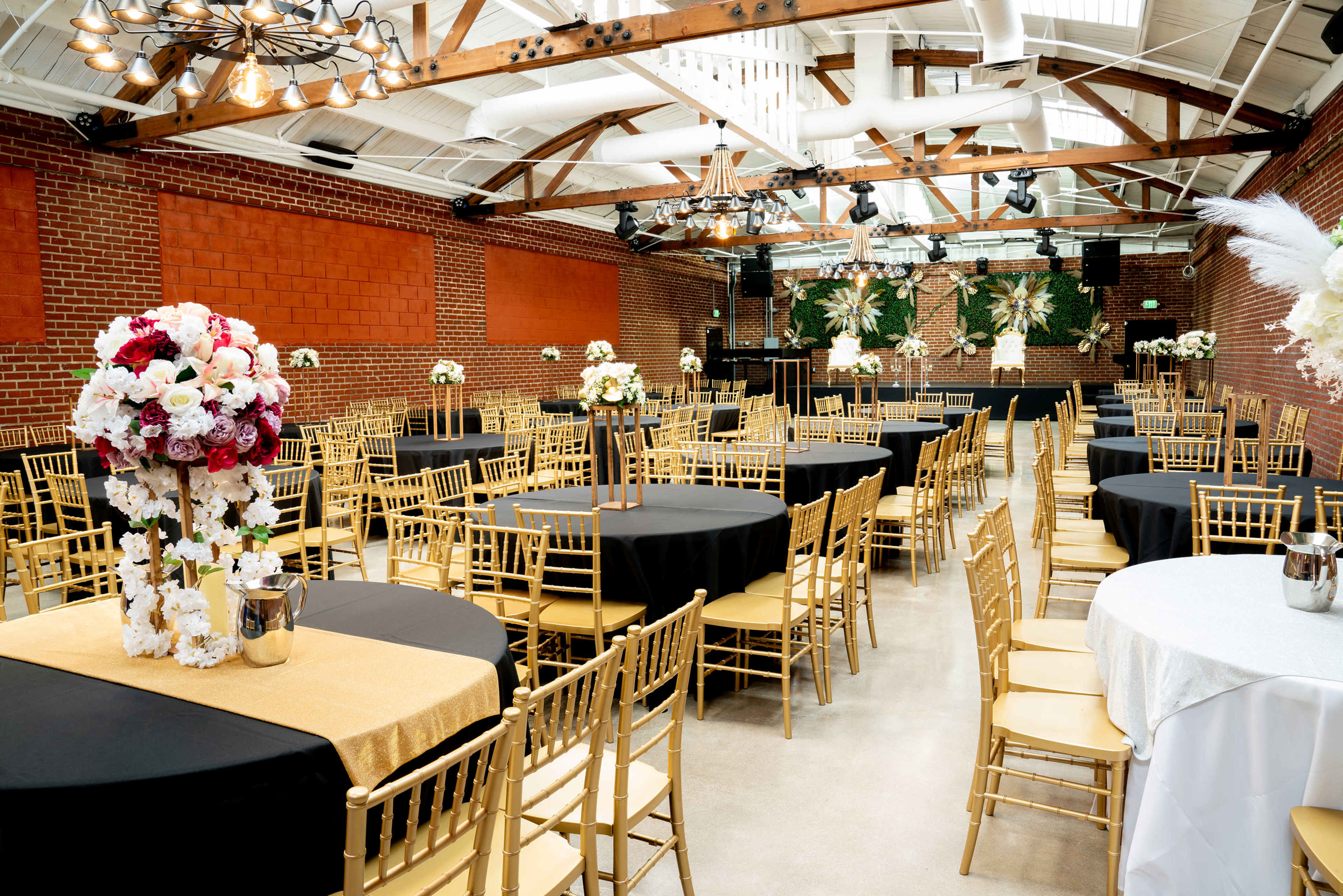 The image shows an elegantly arranged event space with rows of round tables covered in black and gold tablecloths, surrounded by gold chiavari chairs and decorated with floral centerpieces.
