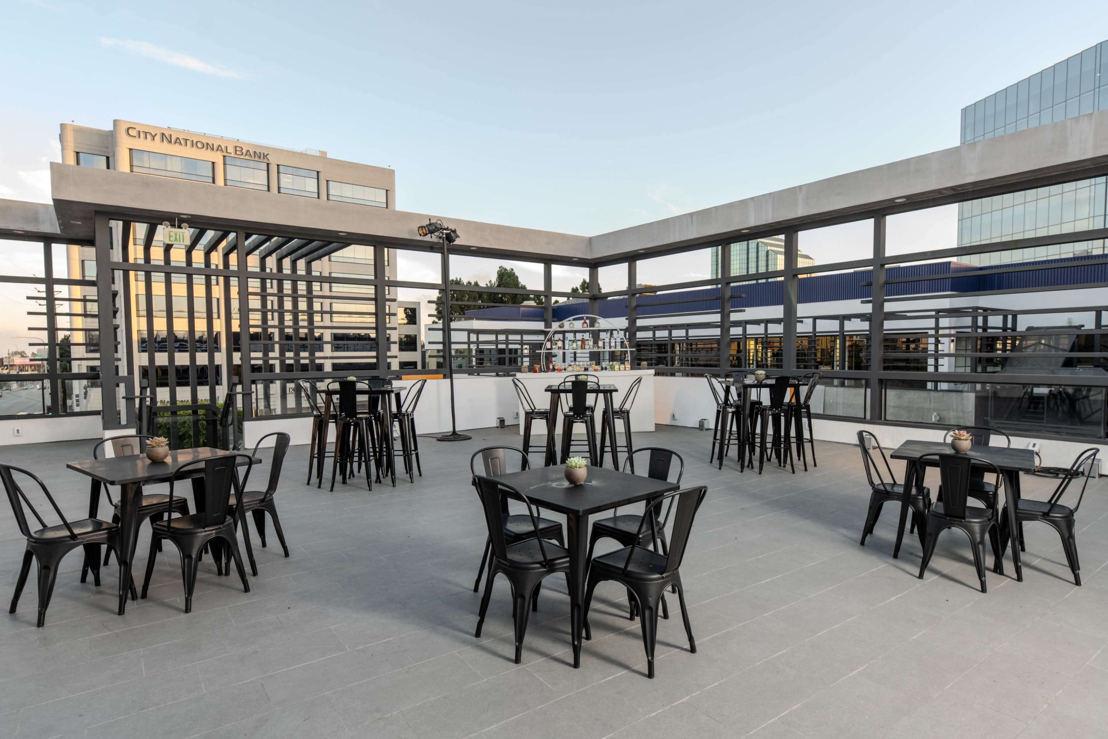 The image depicts a spacious rooftop terrace with several black tables and chairs arranged neatly against a backdrop of buildings.