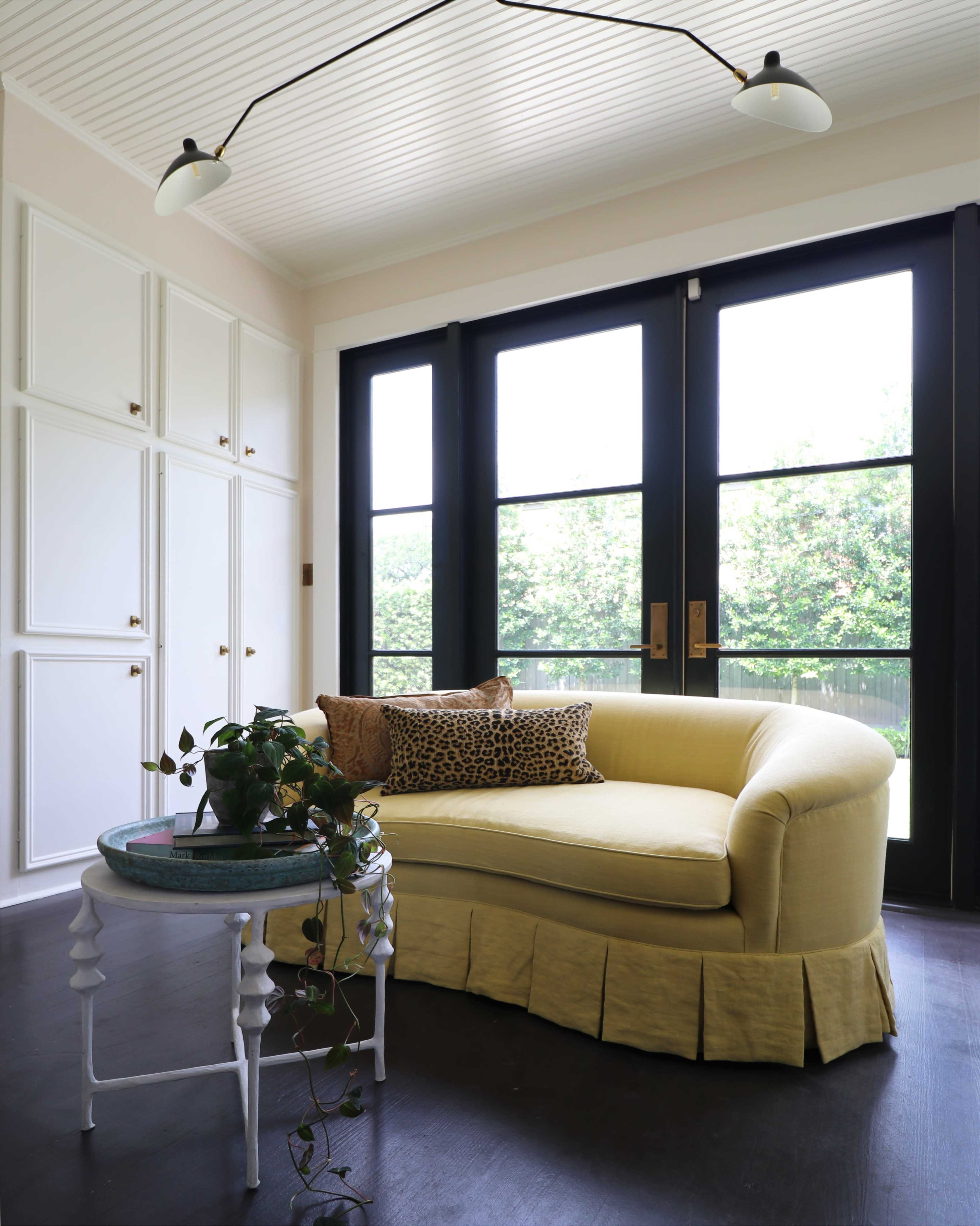 A light yellow curved sofa with a leopard-print pillow sits beside a small round table and a plant near large black-framed windows.