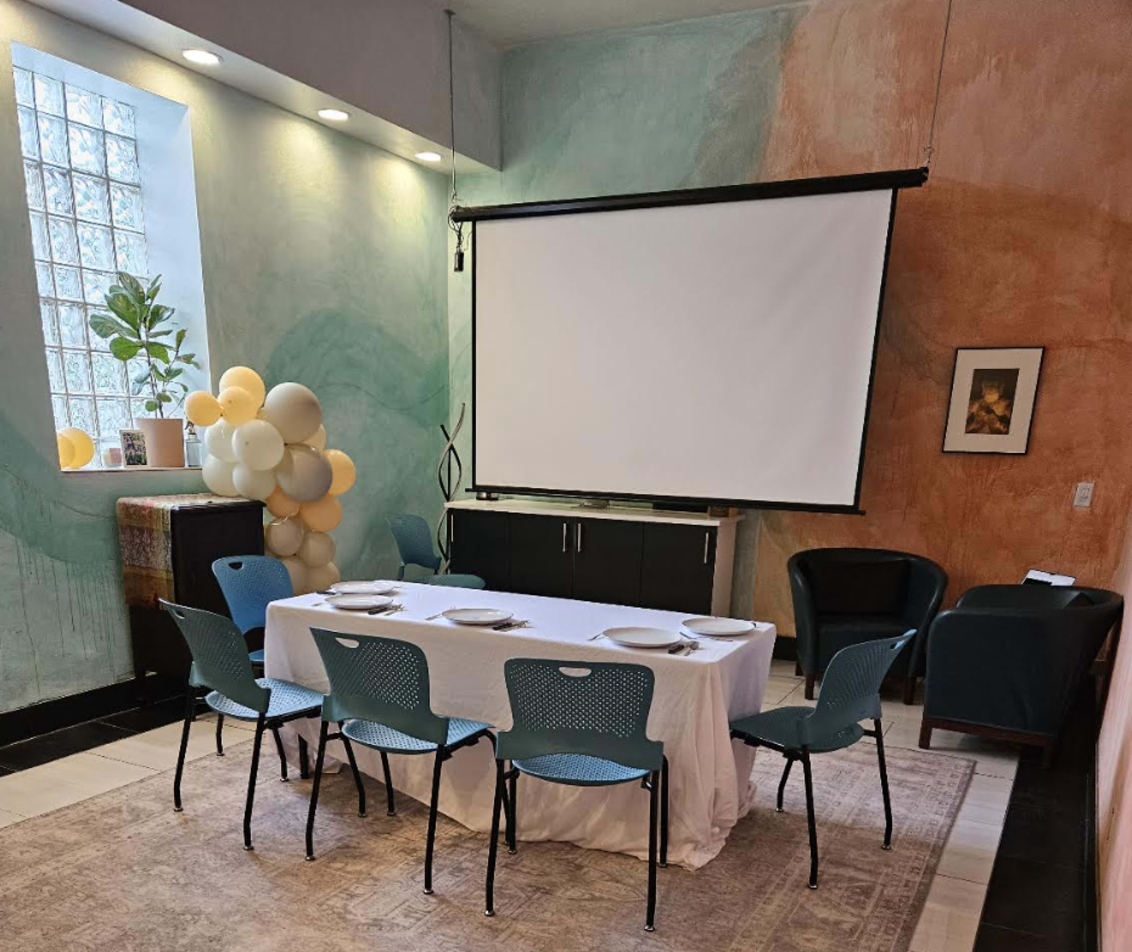 The image shows a dining setup with a white tablecloth, eight blue chairs arranged around a table, and a projector screen mounted on the wall in a brightly colored room.