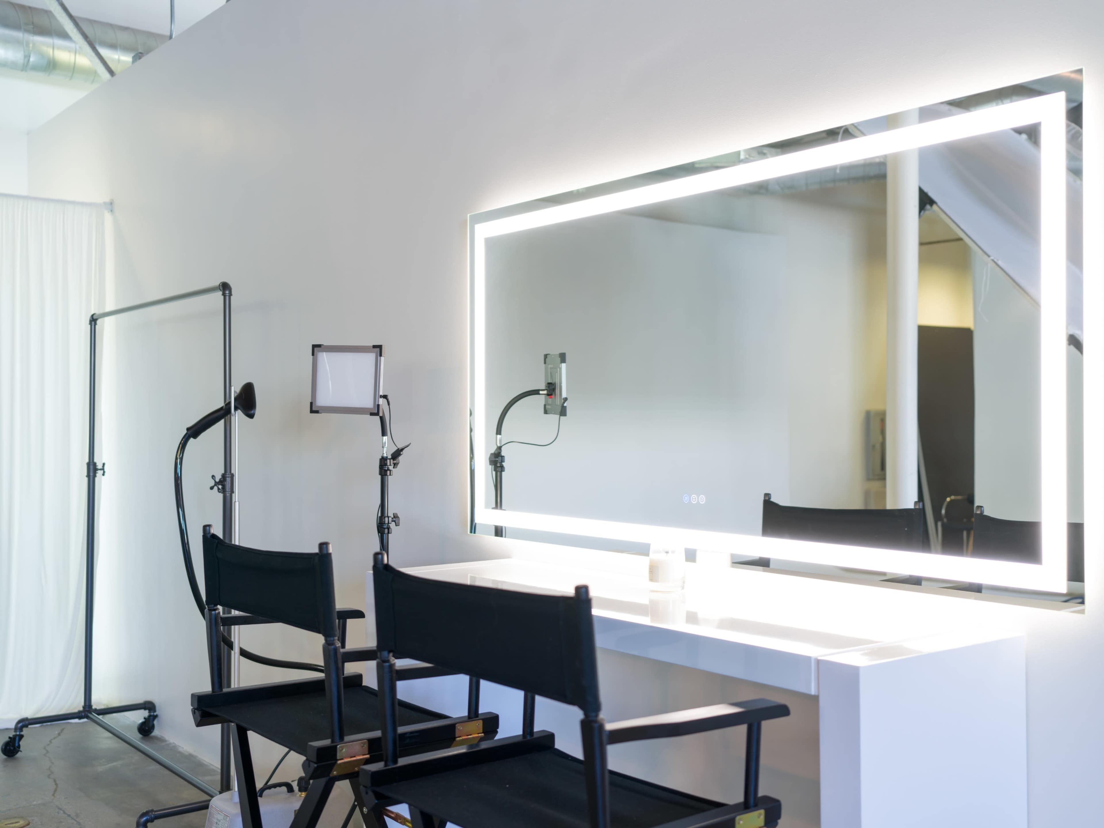 A minimalist makeup studio featuring a large illuminated mirror, two director's chairs, and a standing lamp organized against a plain wall.