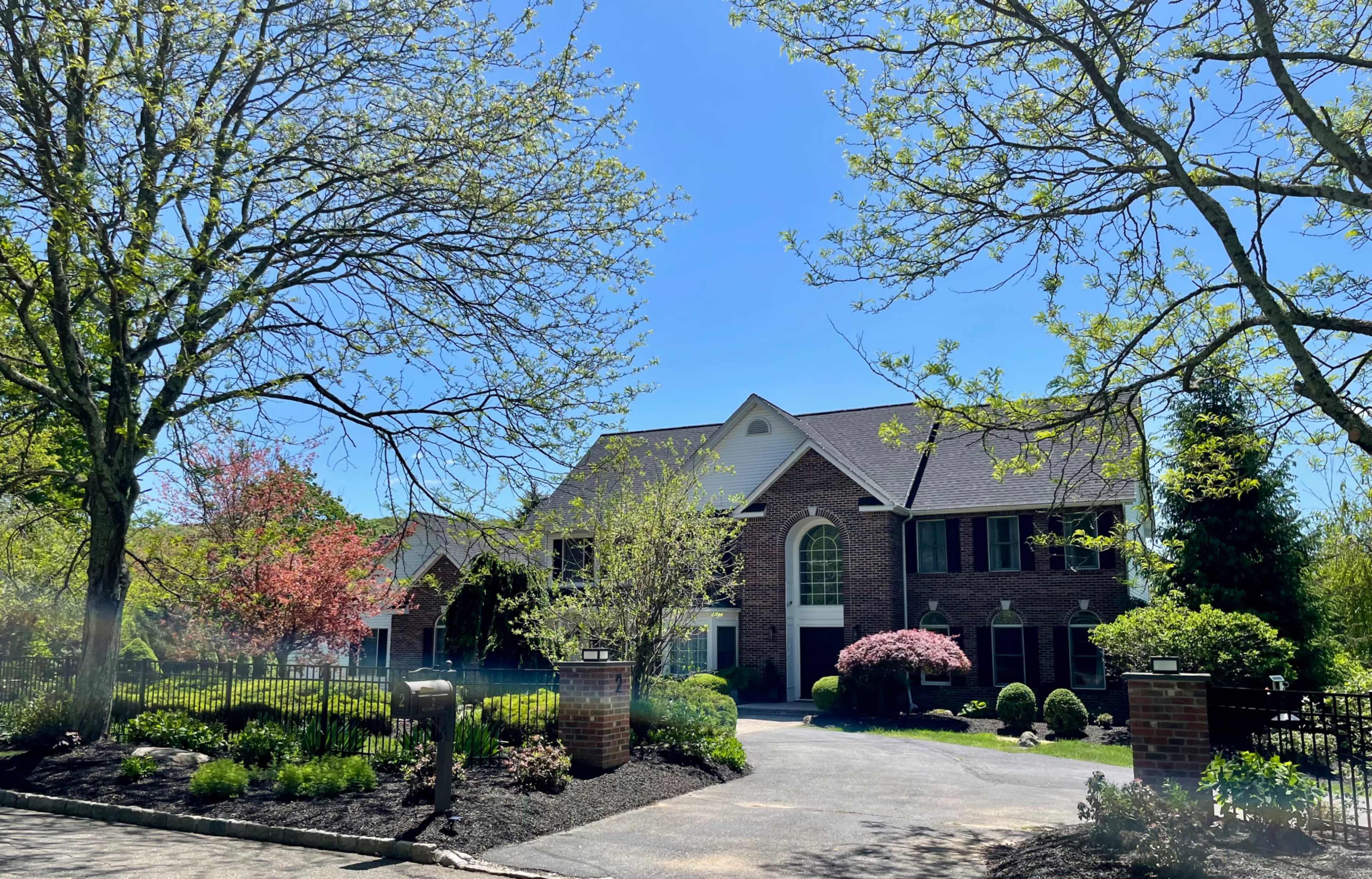 A large brick house with white trim is surrounded by landscaped gardens and trees under a clear blue sky.