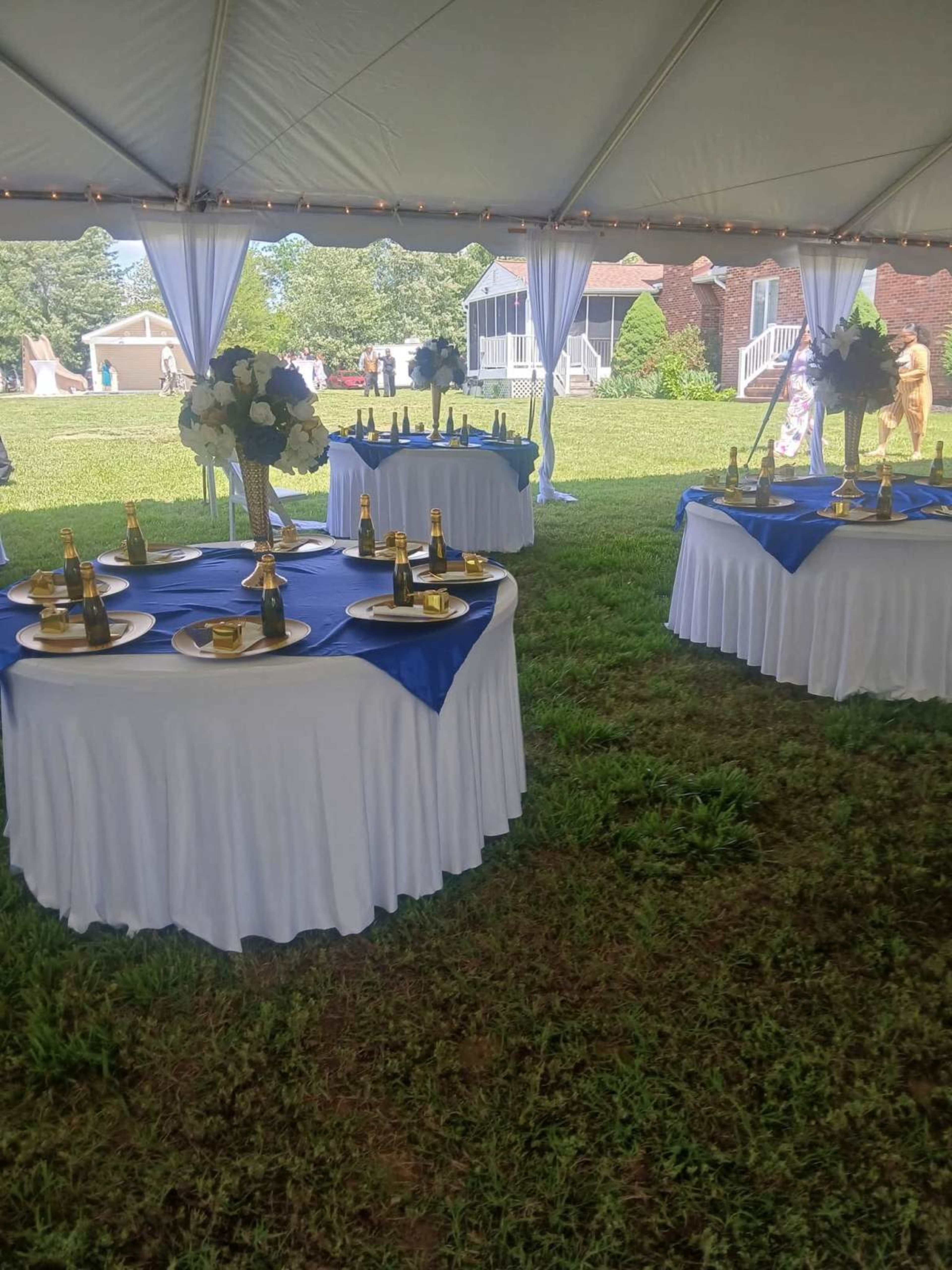 The image shows several round tables adorned with blue tablecloths and gold champagne bottles under a large tent on a grassy area.