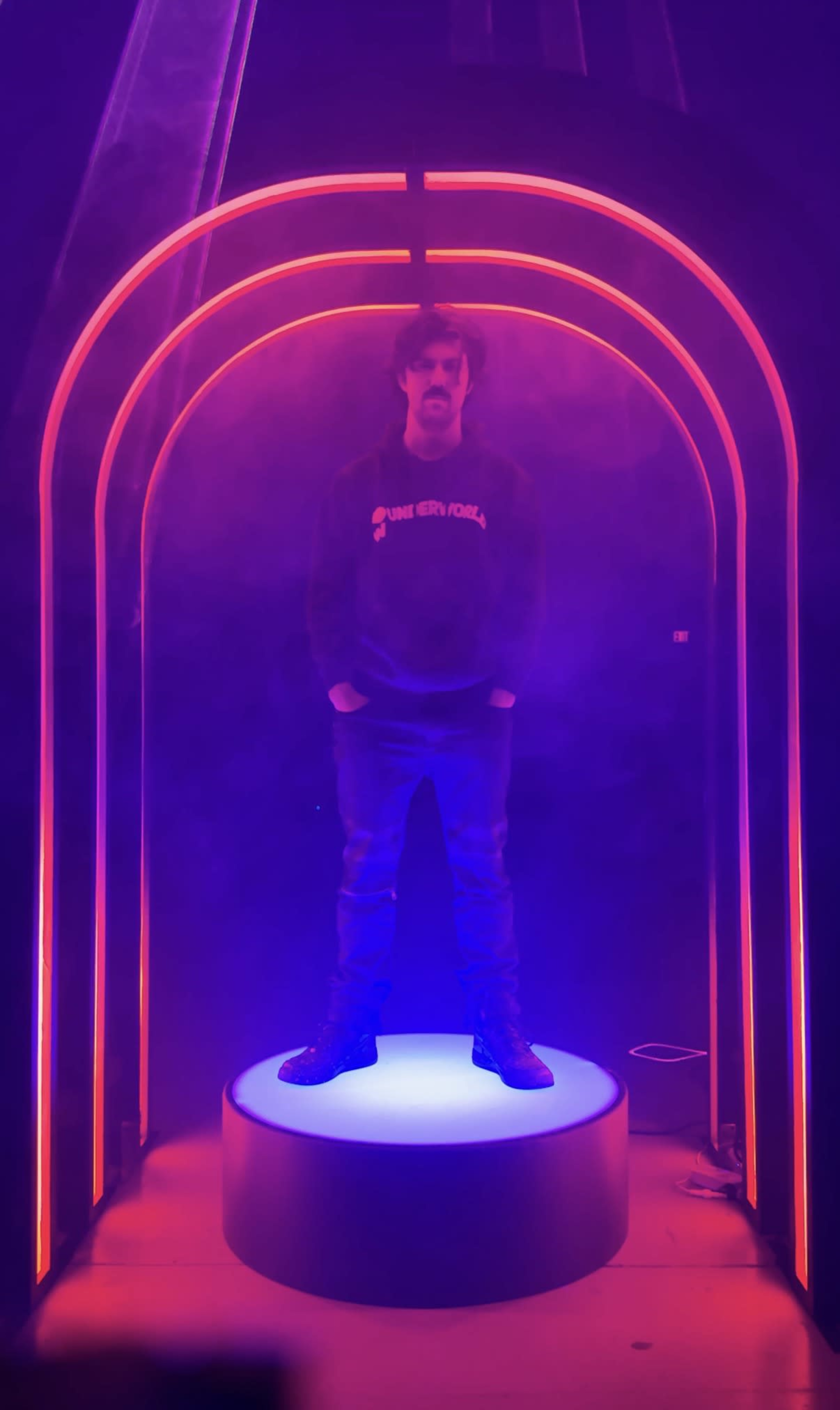 A person stands on a circular platform surrounded by illuminated arches emitting red and purple light in a foggy setting.