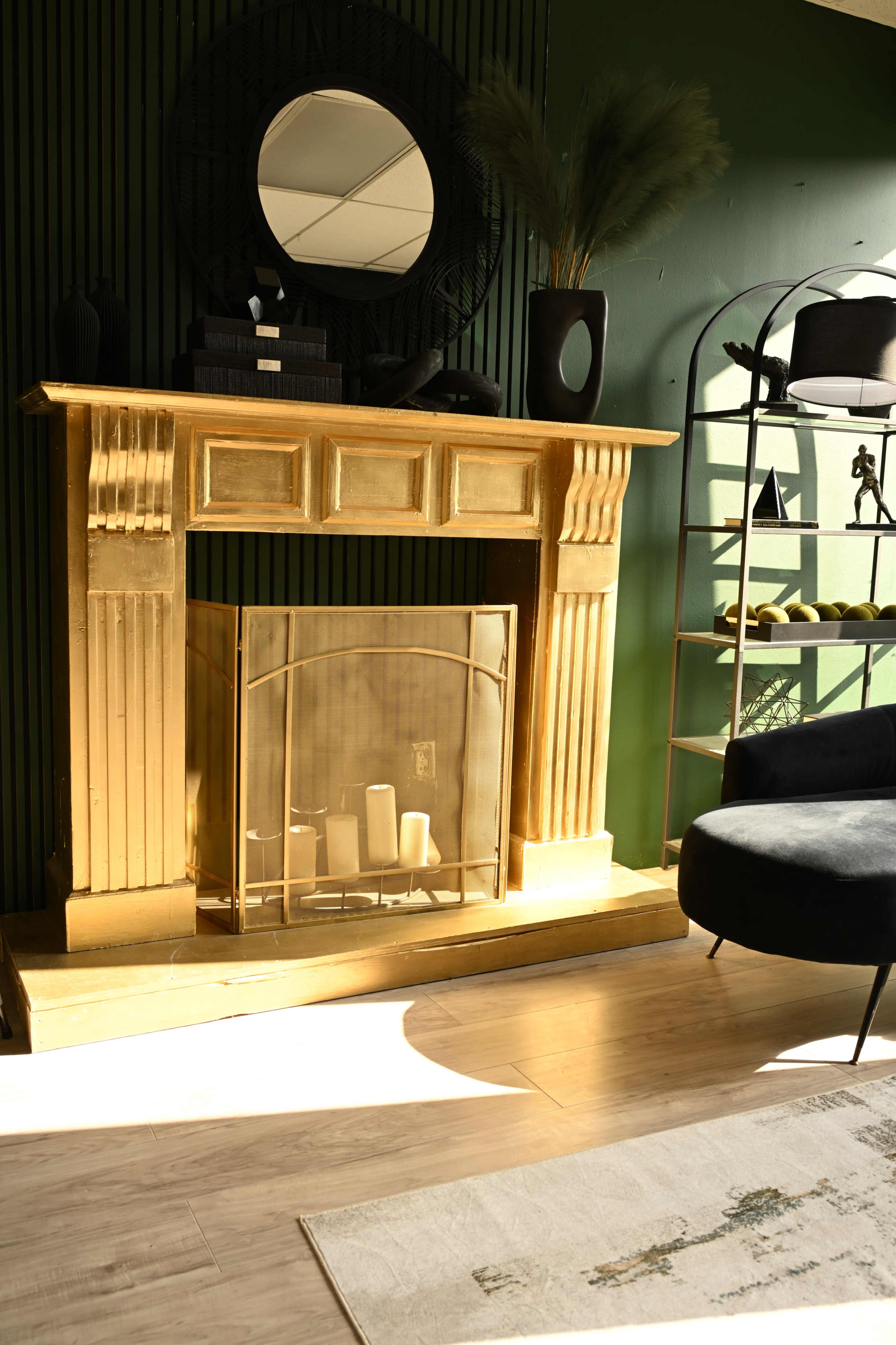 The image features a gold-colored fireplace with a glass screen, positioned against a green wall, alongside a black chair and decorative shelves.