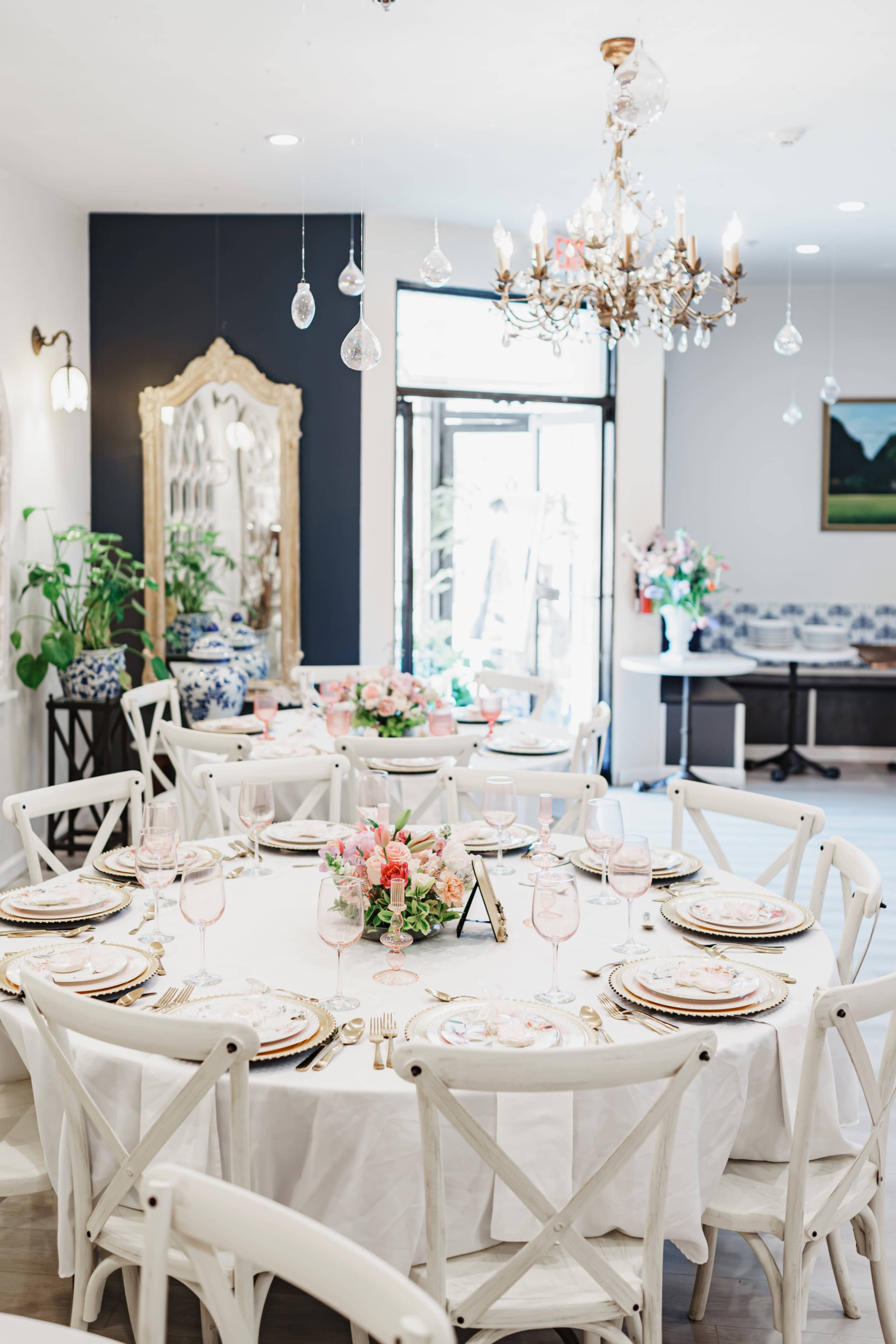A dining room is set up for an elegant event, featuring a round table with a floral centerpiece, fine dishware, and ornate chandelier lighting.