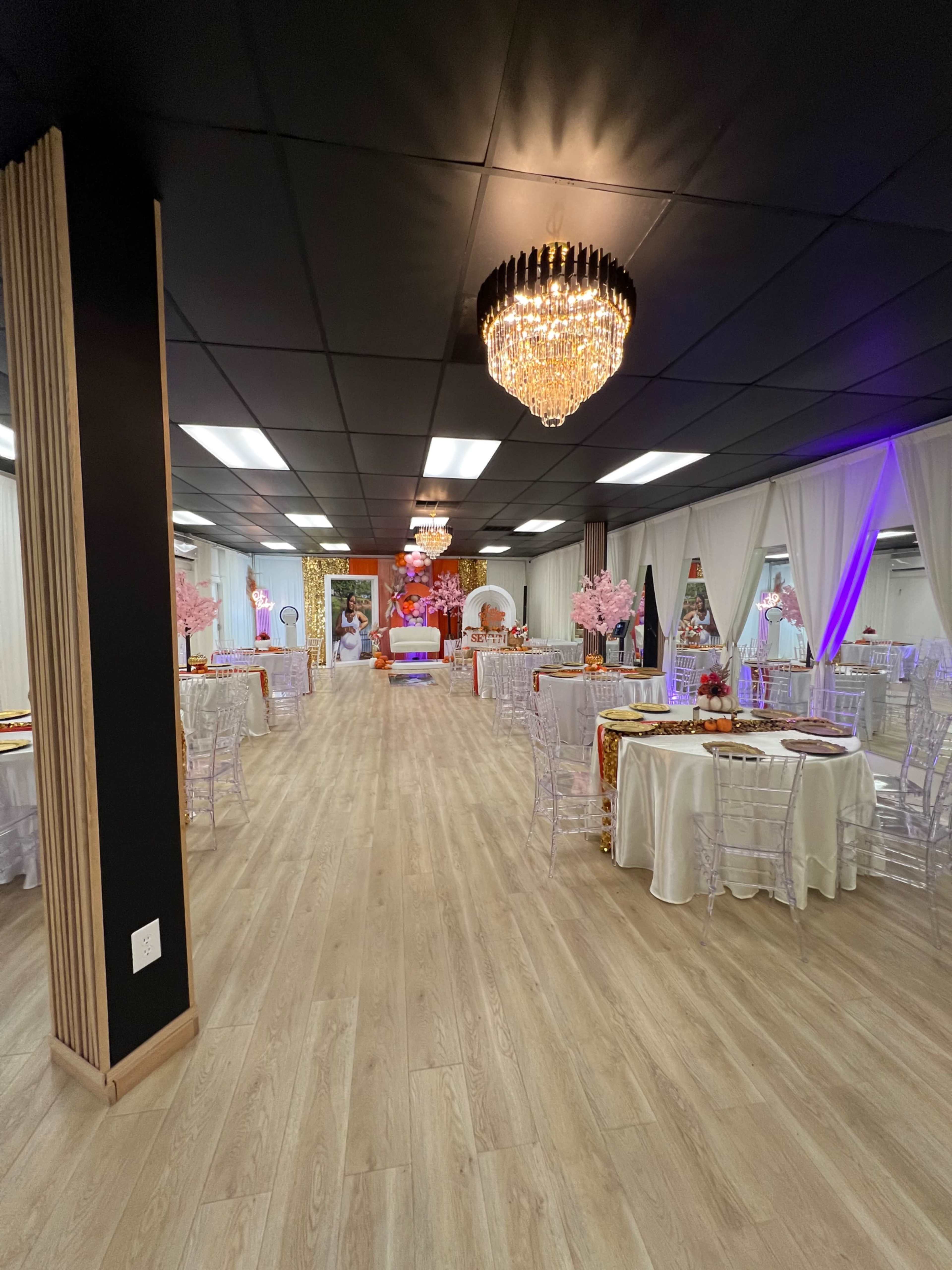 The image shows a spacious event venue with white tables set for dining, surrounded by decorative accents like pink cherry blossom arrangements and elegant lighting fixtures.