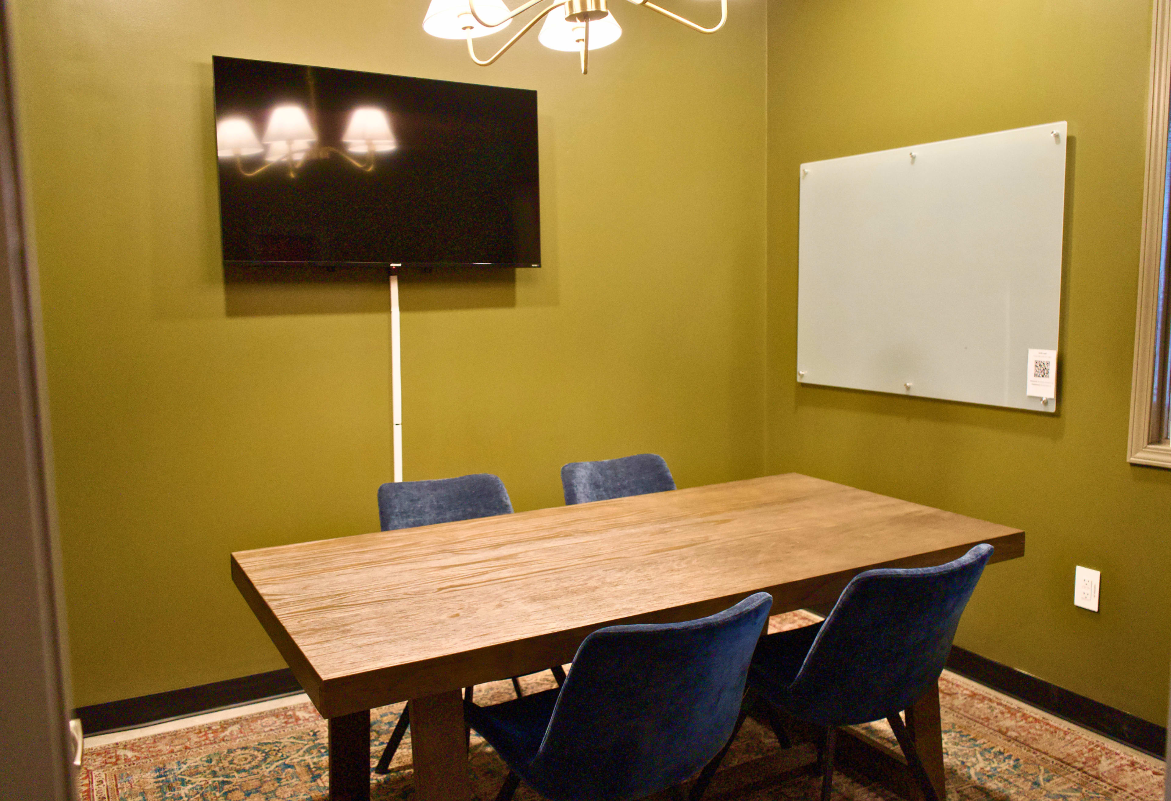 The image shows a small meeting room with a wooden table, four blue chairs, a television mounted on the wall, and a whiteboard.