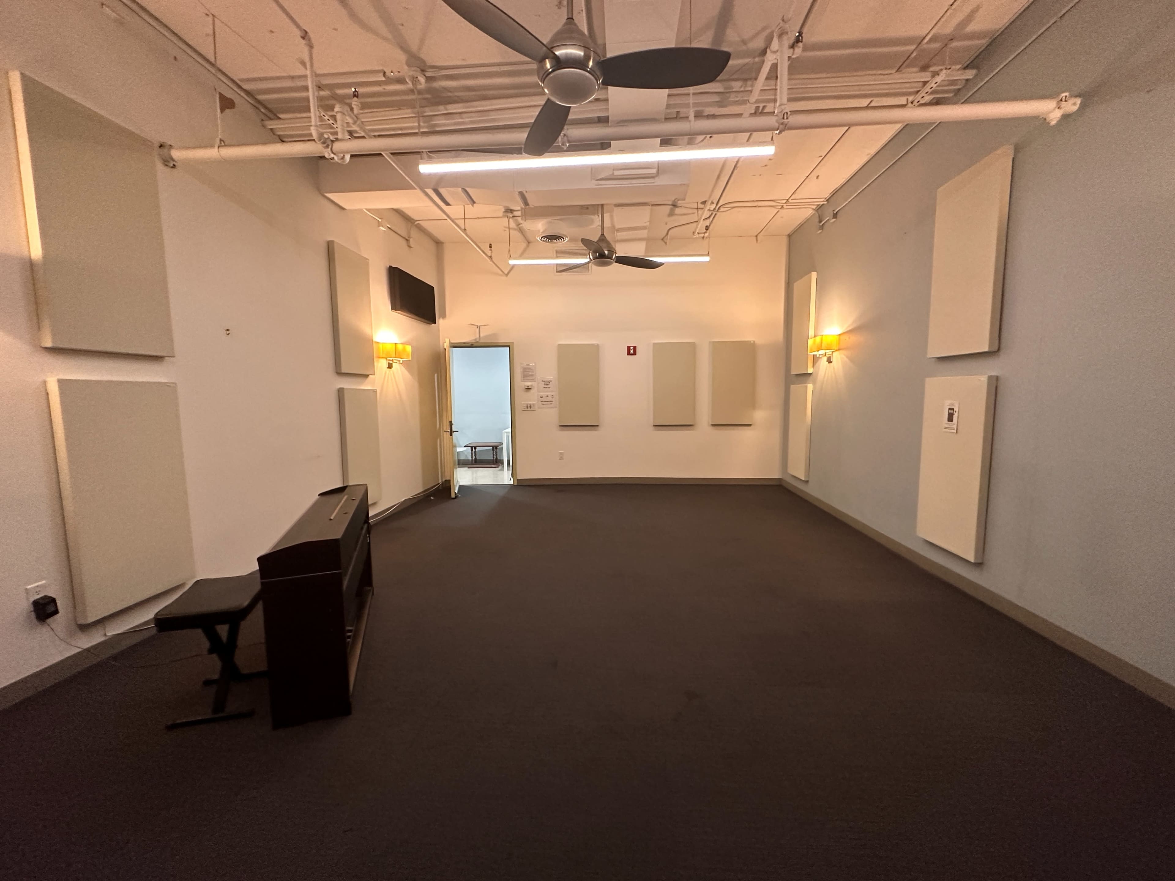 The image shows an empty room with carpeted flooring, wall-mounted soundproof panels, a piano, and ceiling fans.