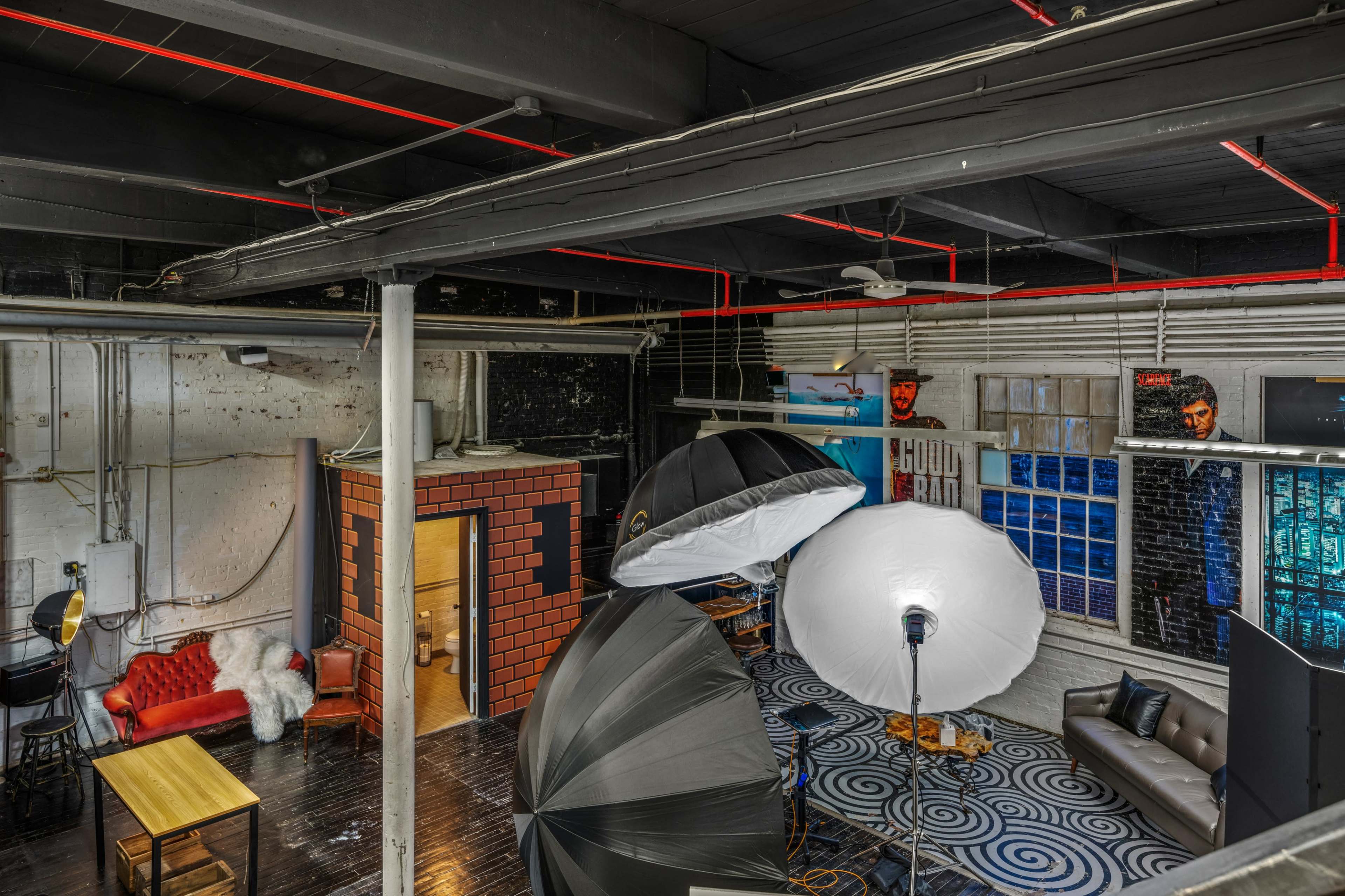 A spacious studio with various photography equipment, including large softbox lights, a red-brick backdrop, and different seating areas.