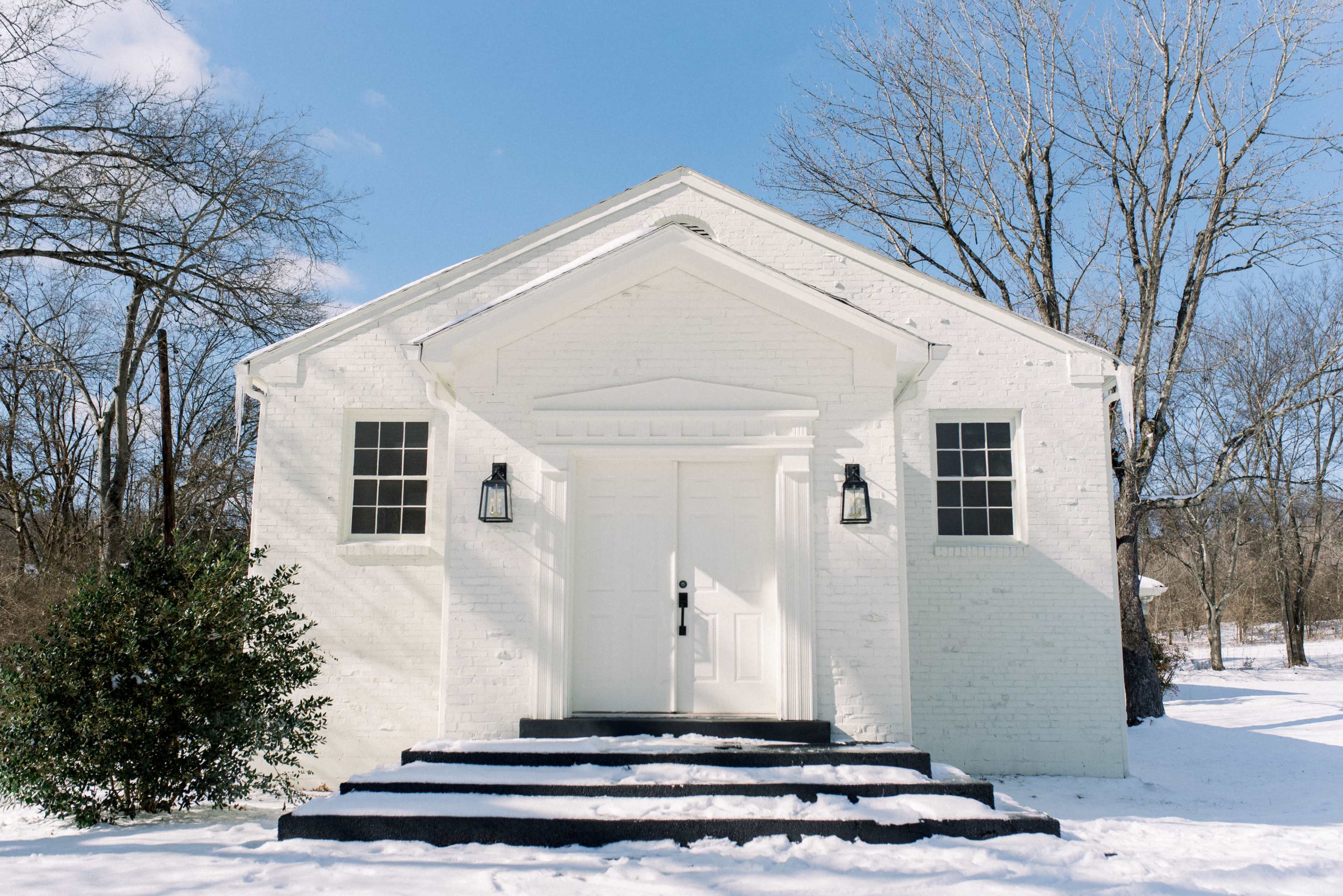 A white brick building with a gabled roof and two front windows stands in a snow-covered landscape.