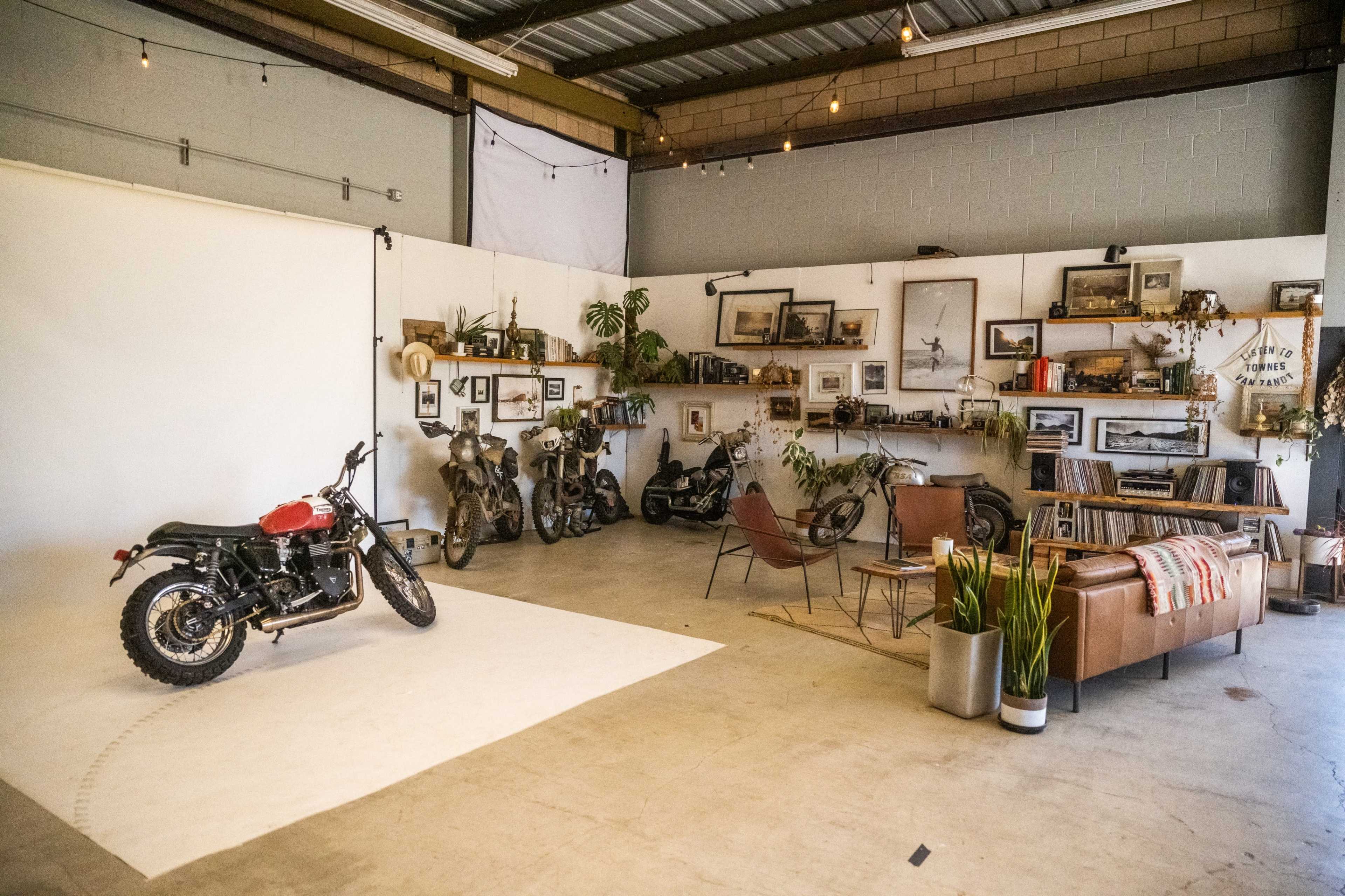 A motorcycle studio features a red motorcycle on a white backdrop, surrounded by vintage motorcycles and a seating area with plants, bookshelves, and framed photographs on the walls.