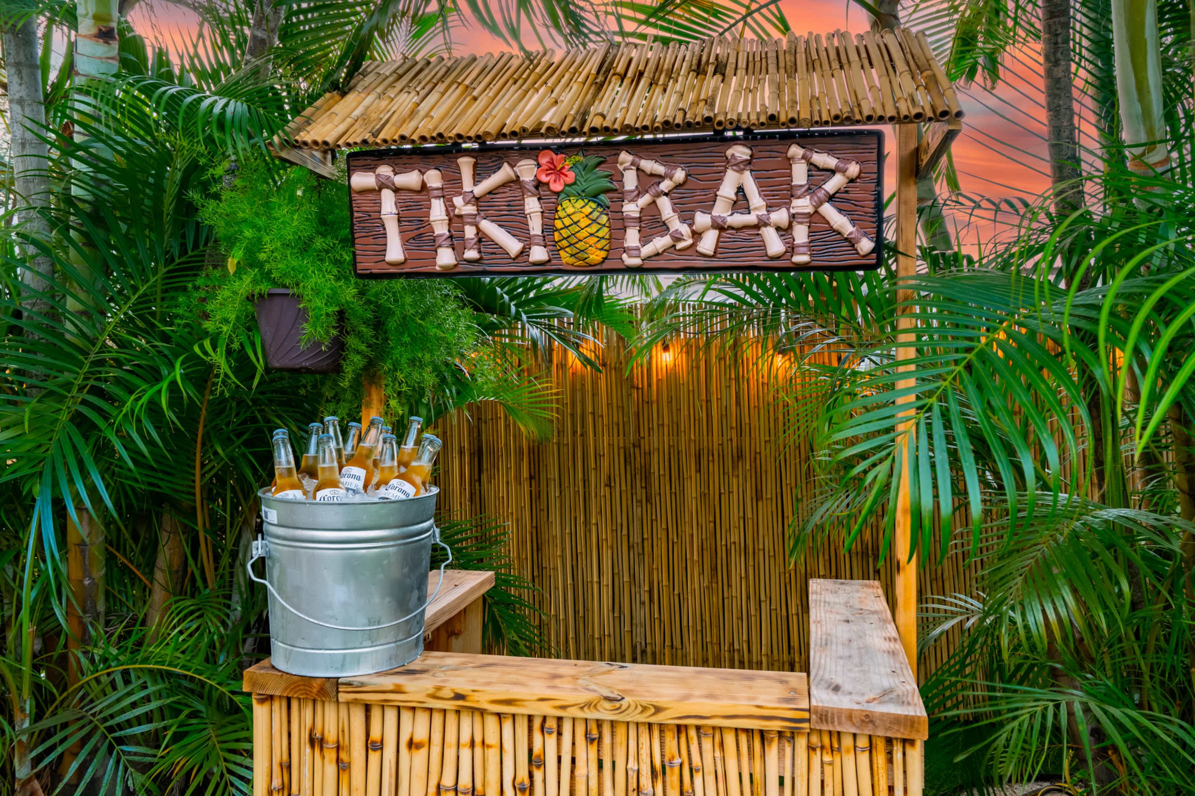 A Tiki bar with a bamboo structure and a sign made of natural materials stands in a lush, tropical setting with a metal bucket of beverages.