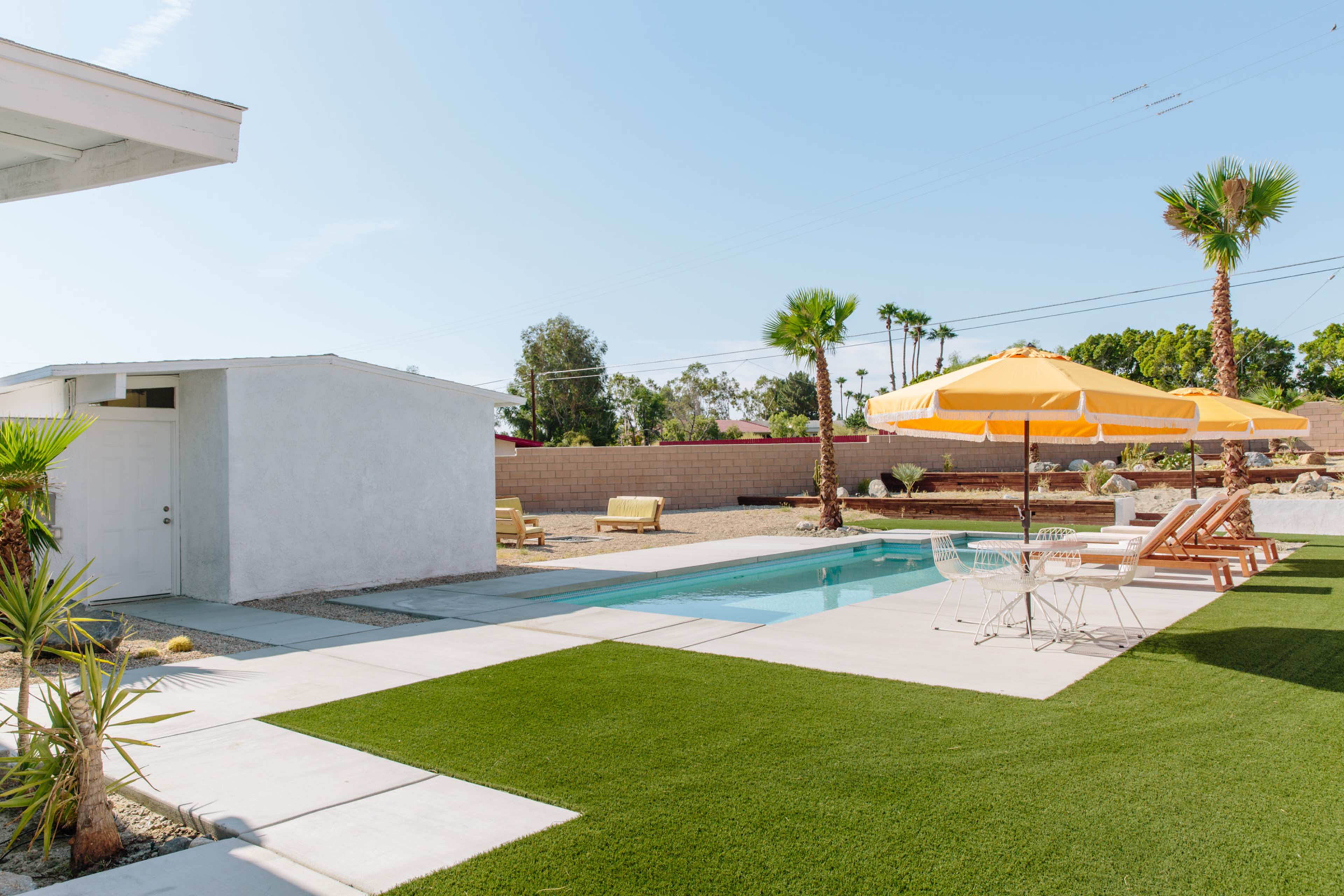 A modern backyard with a swimming pool, lounge chairs, an umbrella, and a small building beside landscaped grass and palm trees.