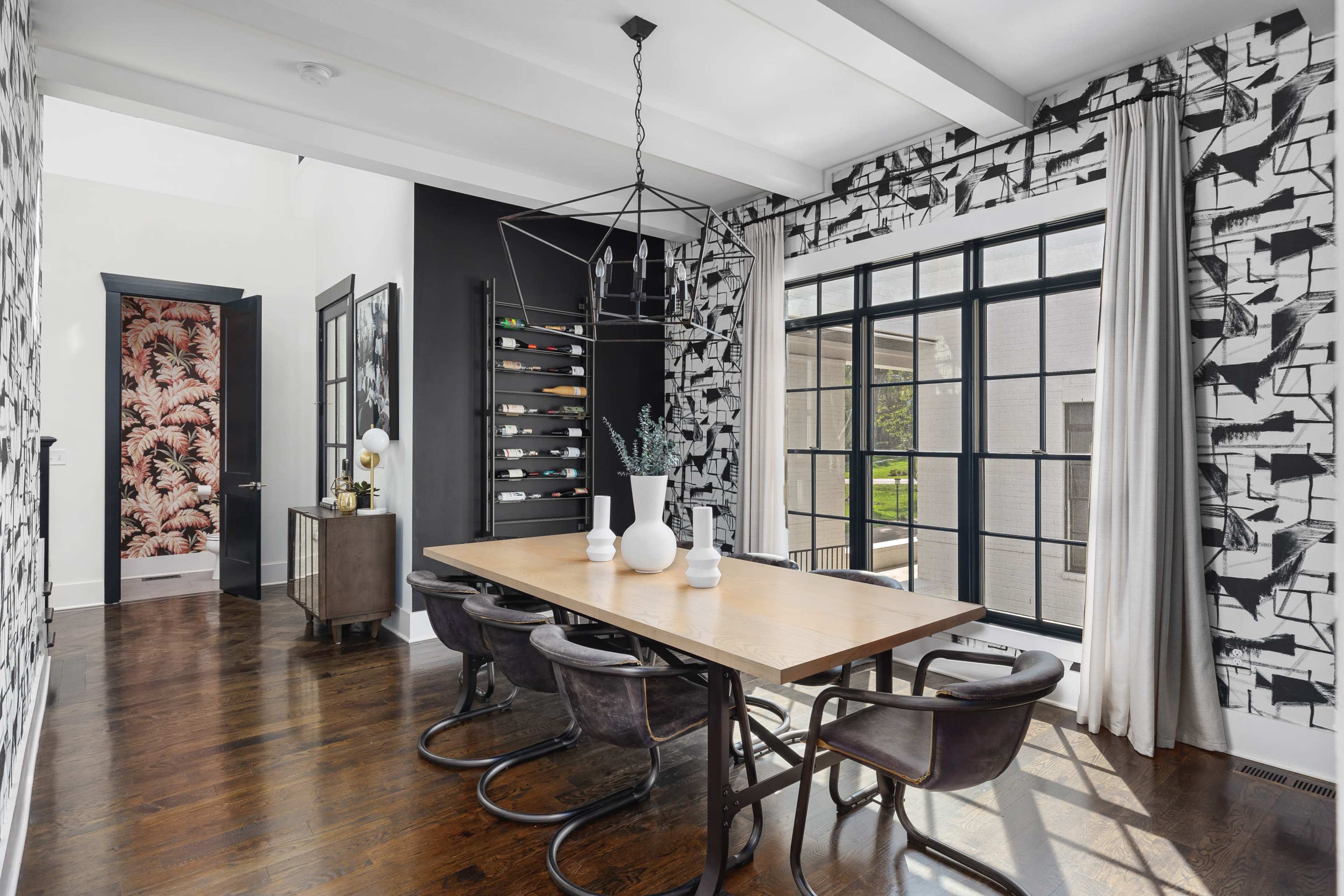 A dining area features a wooden table surrounded by black chairs, with large windows allowing natural light to fill the space, and bold wallpaper adding visual interest to the walls.