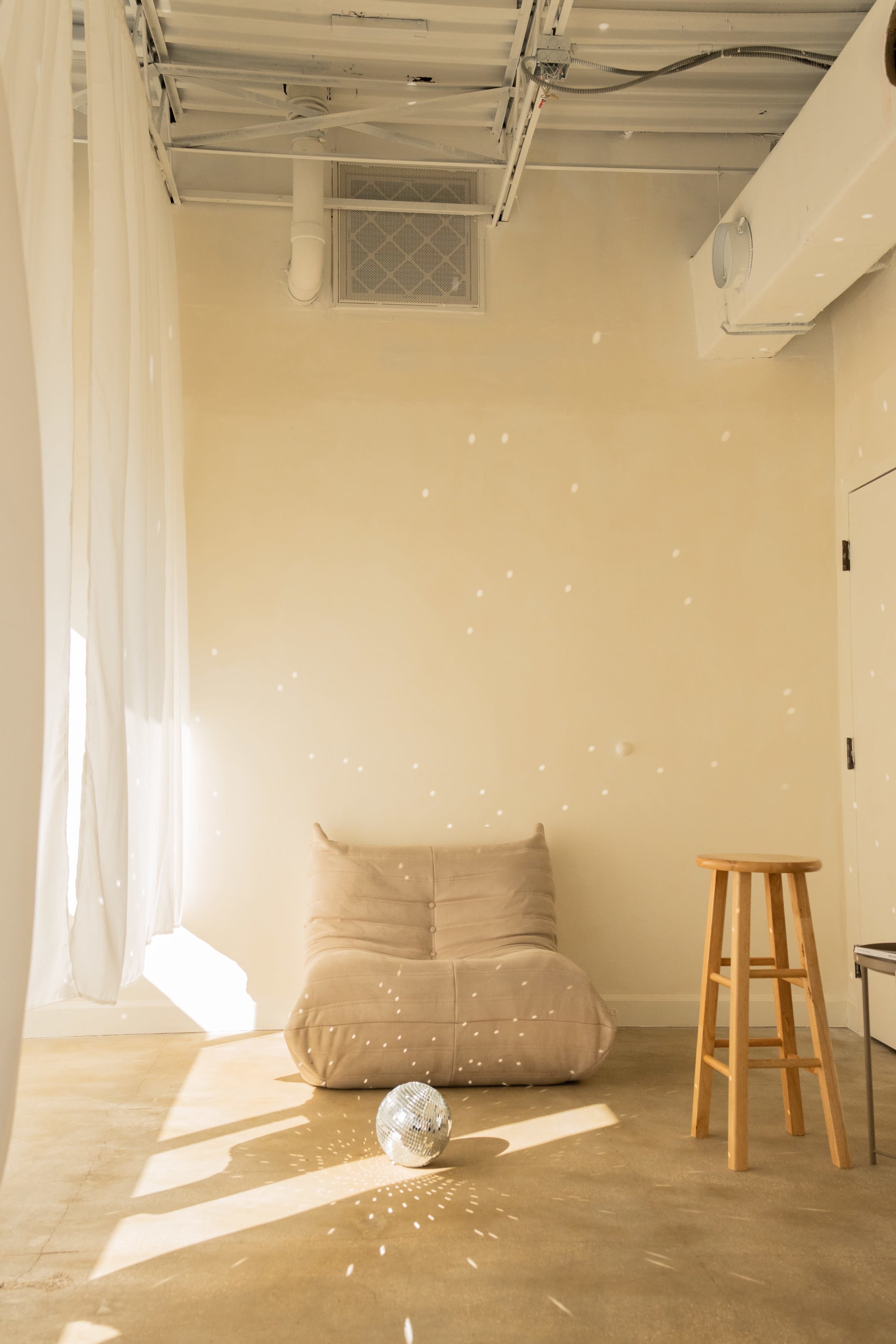 A quiet room features a beige floor chair and a wooden stool, with sunlight casting dots on the walls from a disco ball on the floor.