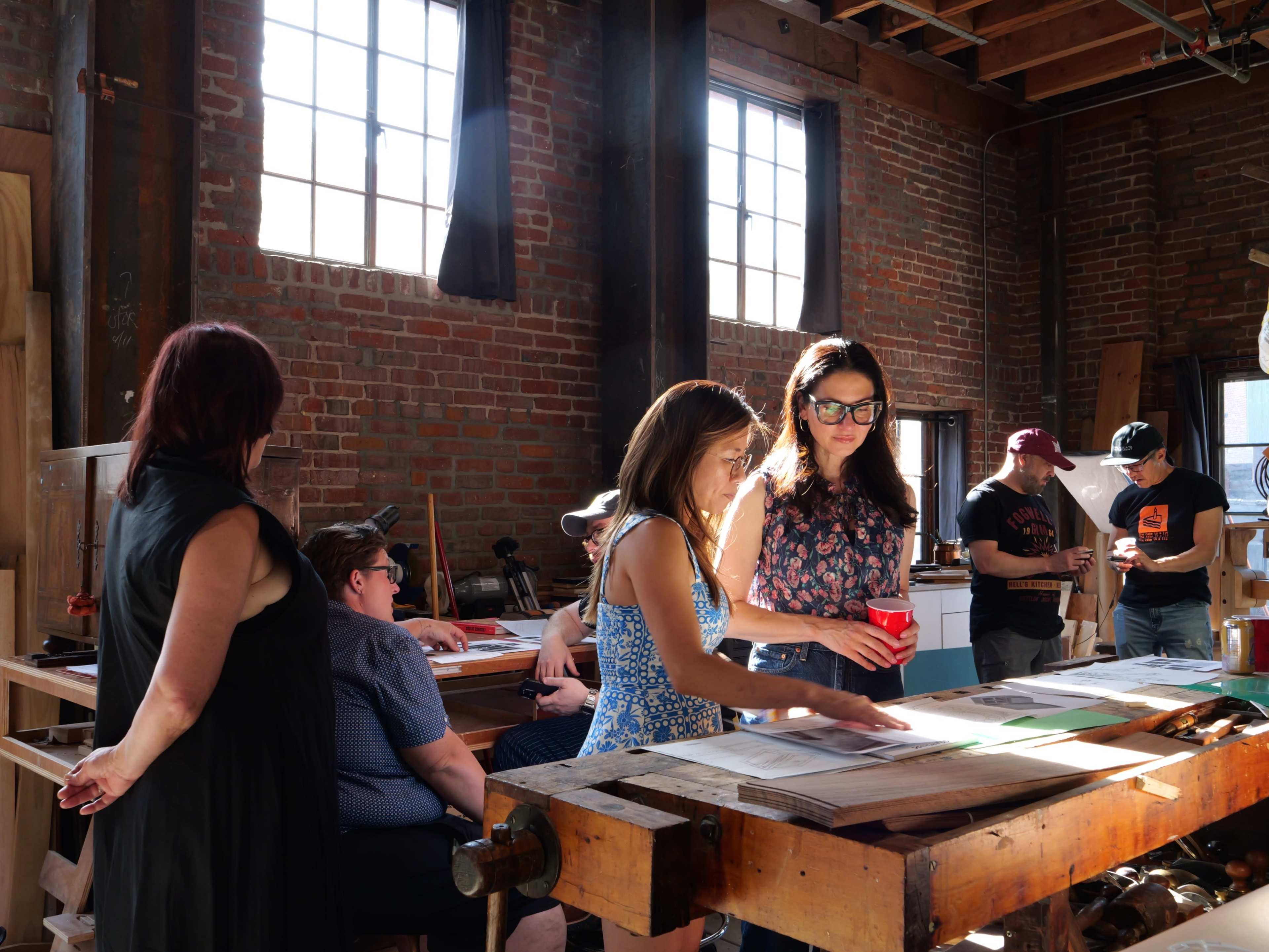 A group of people examines printed materials around a large wooden table in a well-lit workshop with brick walls and large windows.