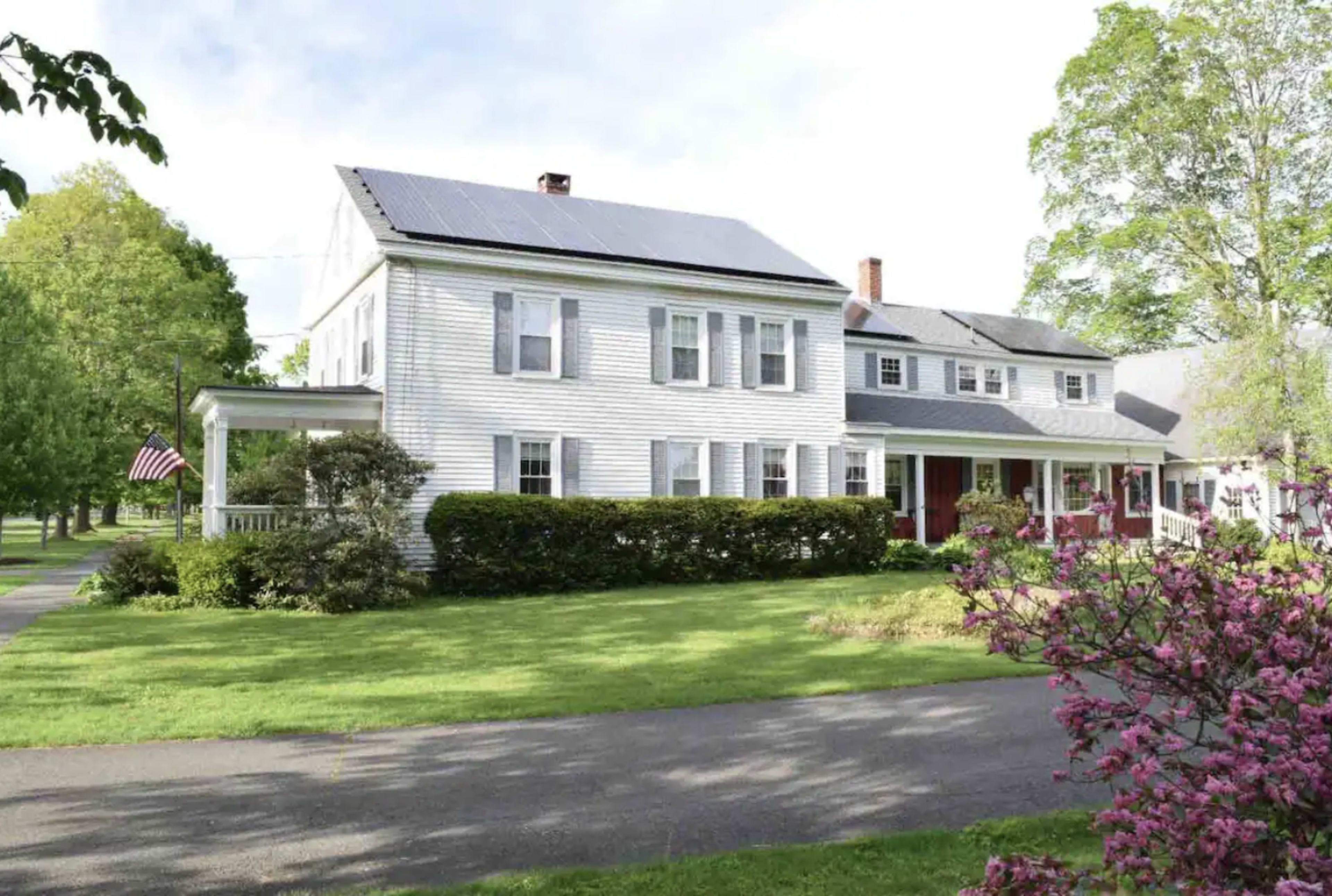 A white two-story house with solar panels on the roof, surrounded by green lawns and trees, with a porch and American flag in front.