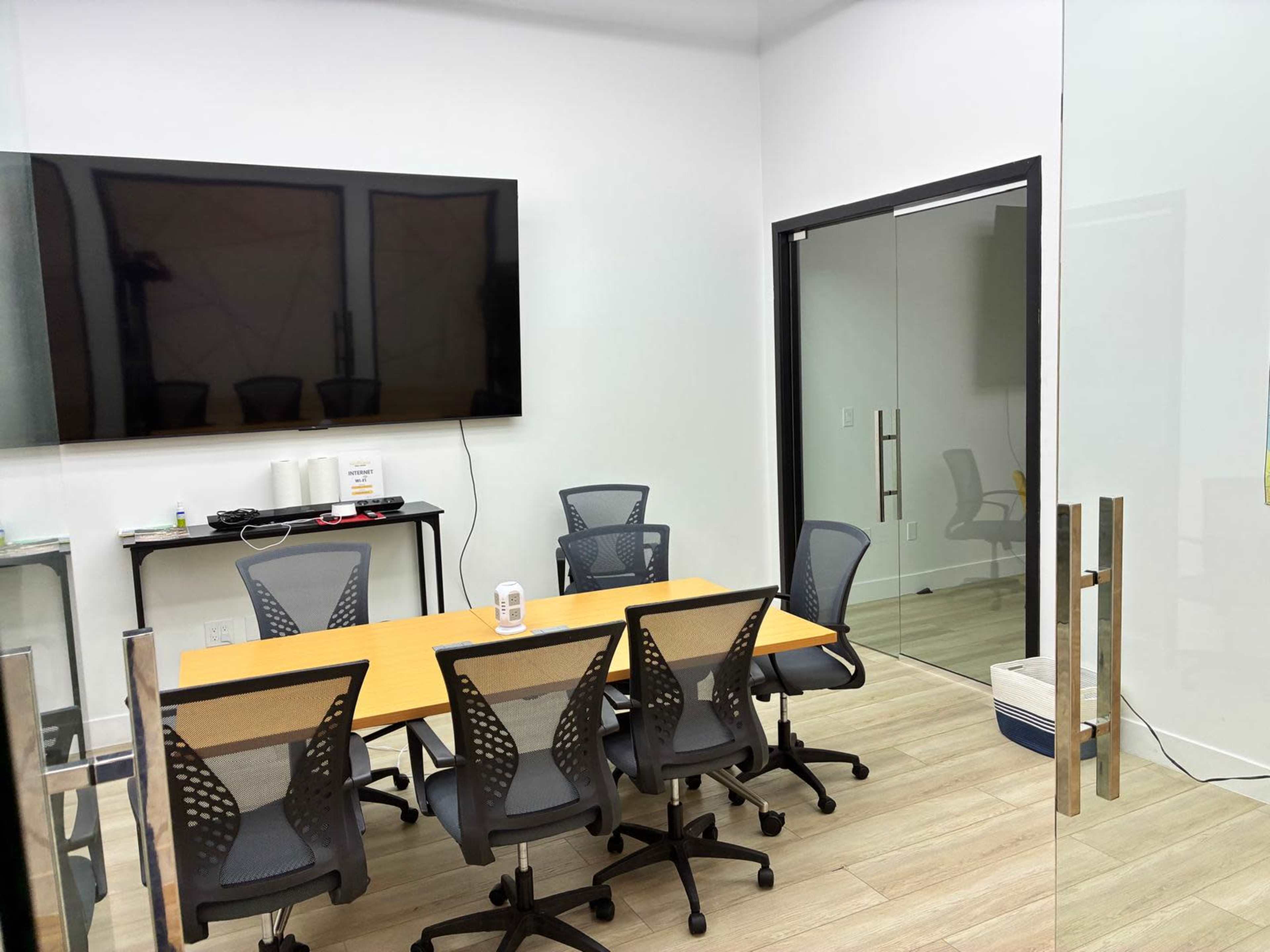 The image shows a modern conference room with a large wall-mounted screen, a long table surrounded by six office chairs, and a glass door.