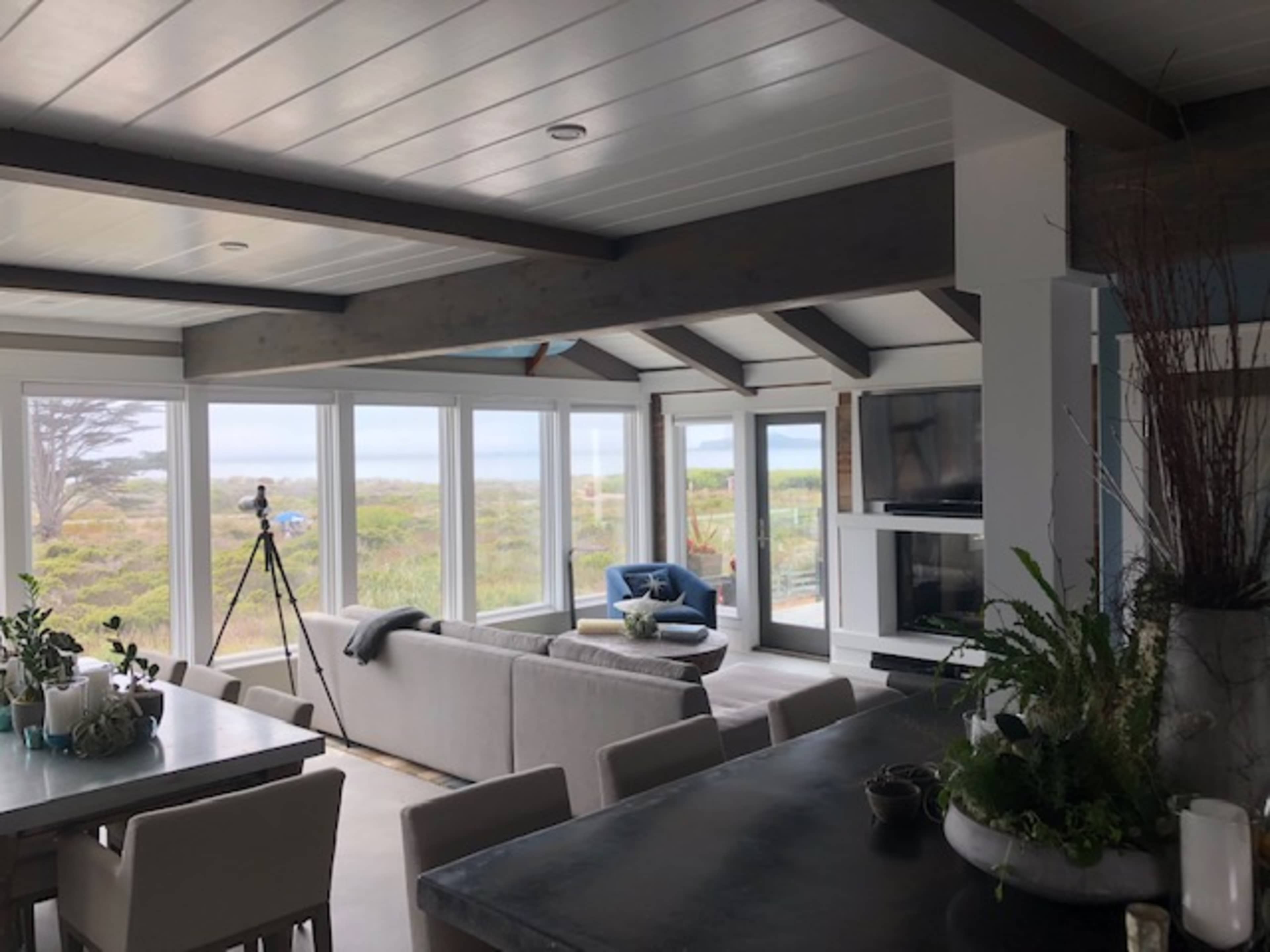 A modern living area with large windows overlooking a coastal landscape, featuring a gray sofa, a television, and a dining table.