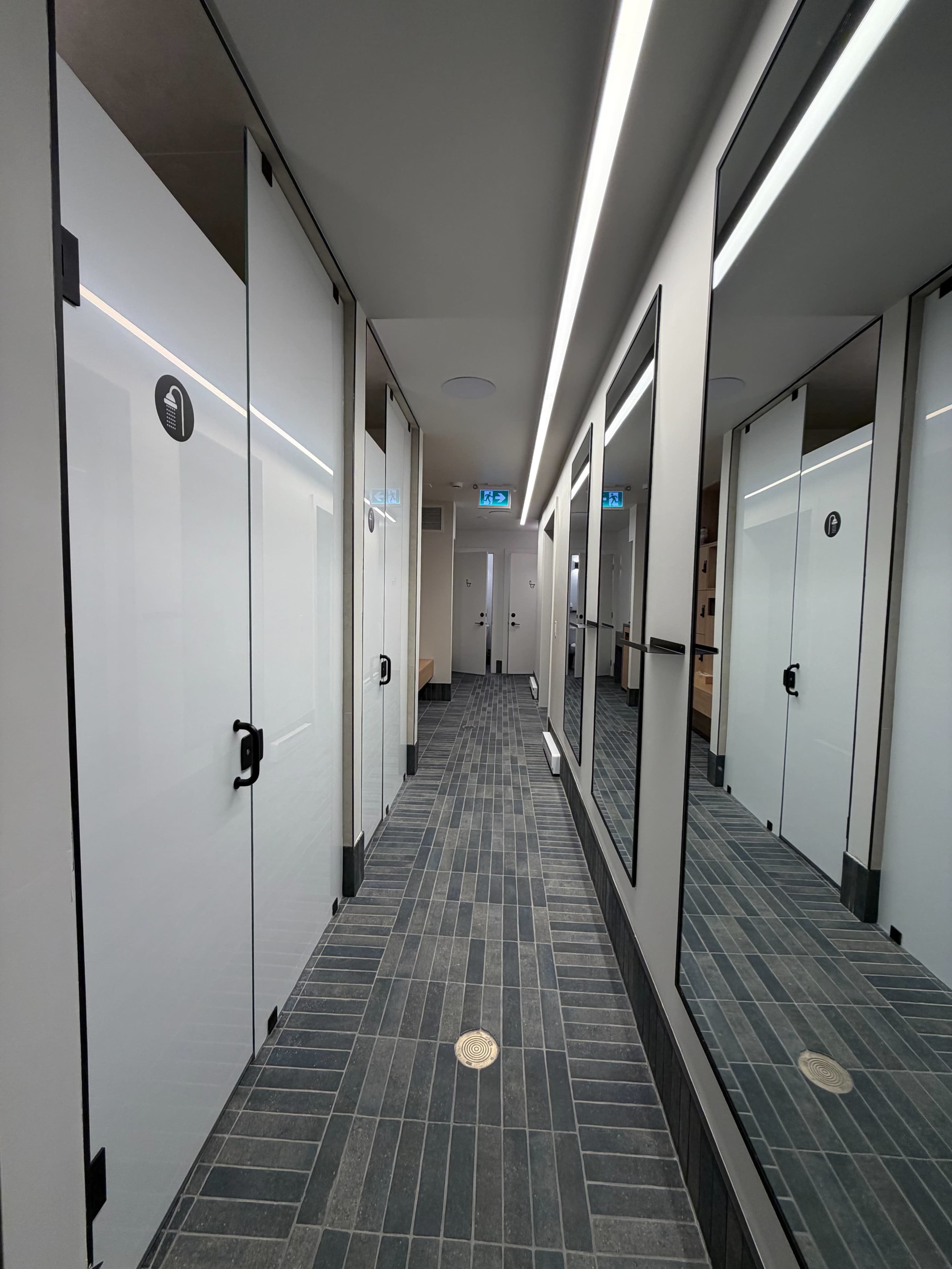 The image shows a modern restroom hallway with multiple frosted glass stall doors and large mirrors along the walls.