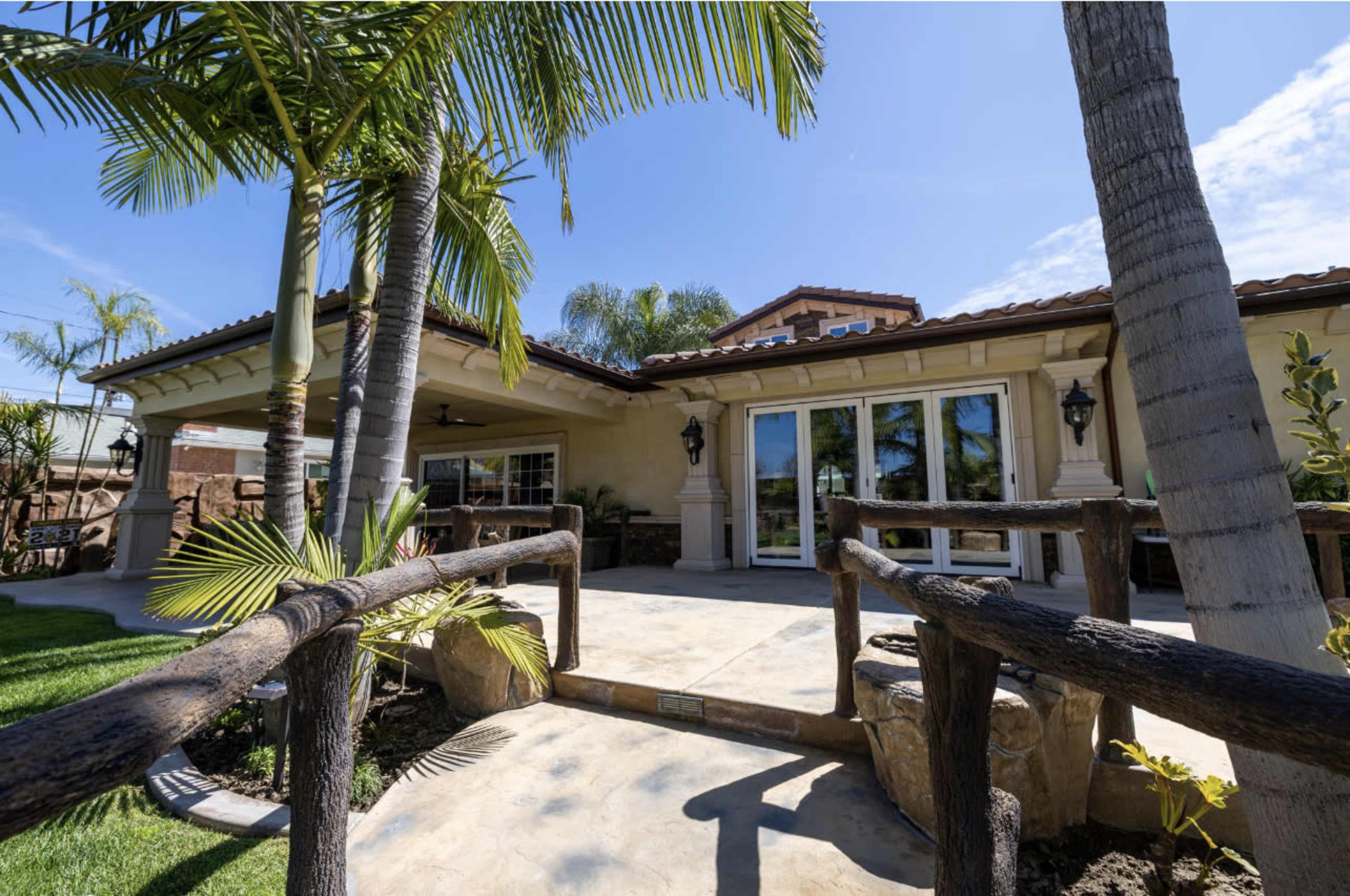 A backyard area with a covered patio, palm trees, and a view of a house featuring large windows.
