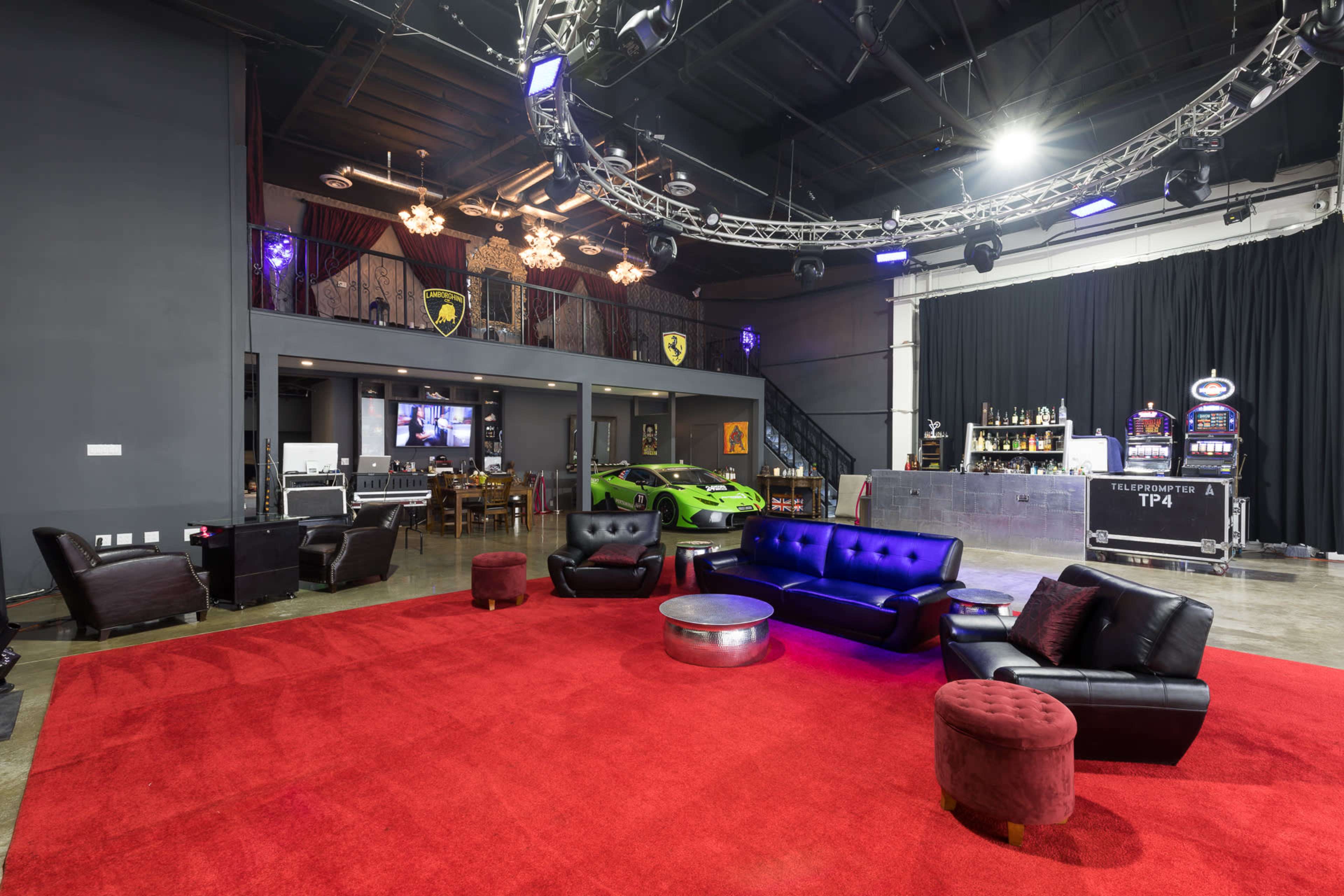 The image shows a spacious entertainment area featuring a red carpet, black leather couches, a bar, a staircase, and a green sports car in the background.