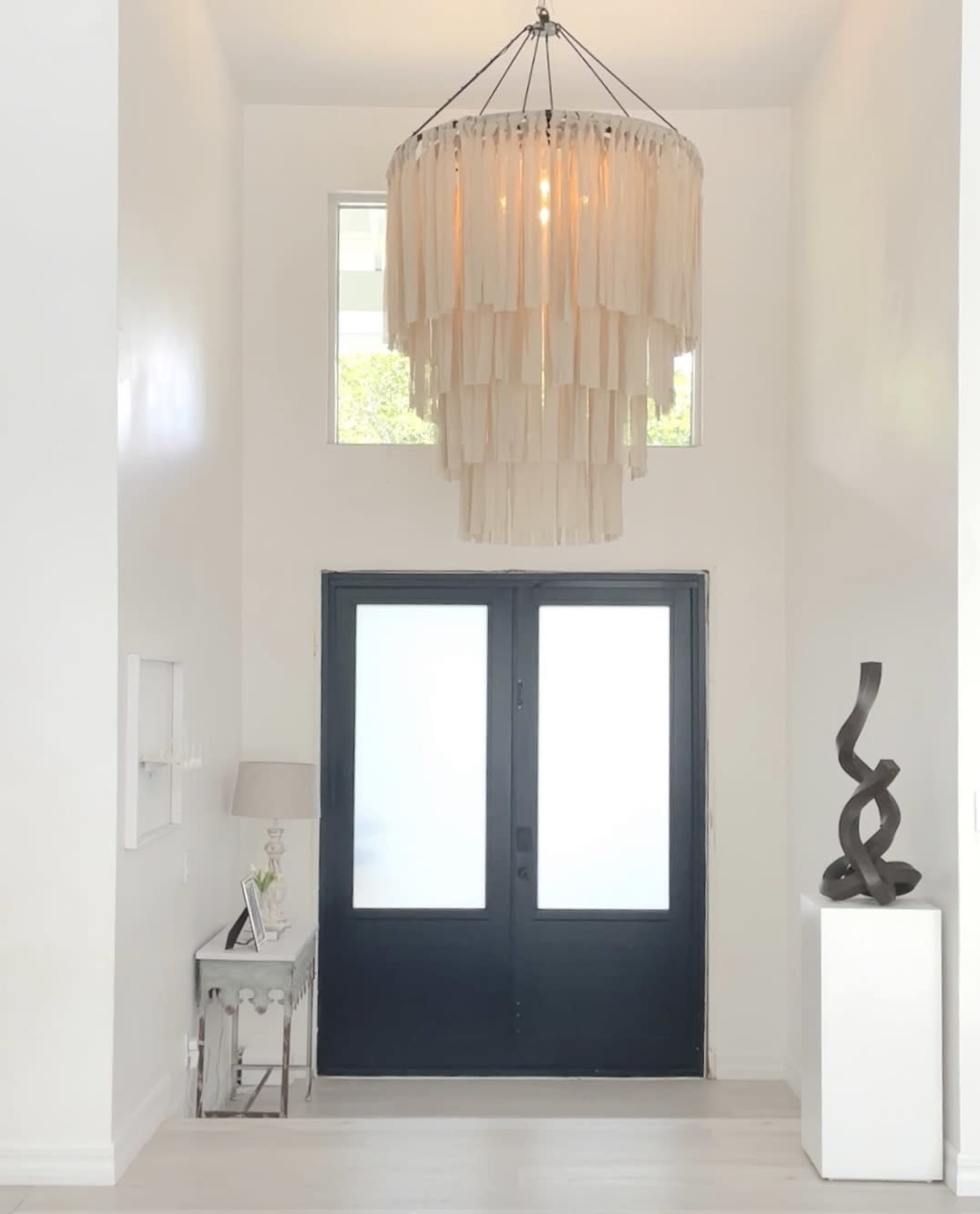 A well-lit entryway featuring a large, fringed chandelier above a pair of black double doors and a modern sculpture on a pedestal.
