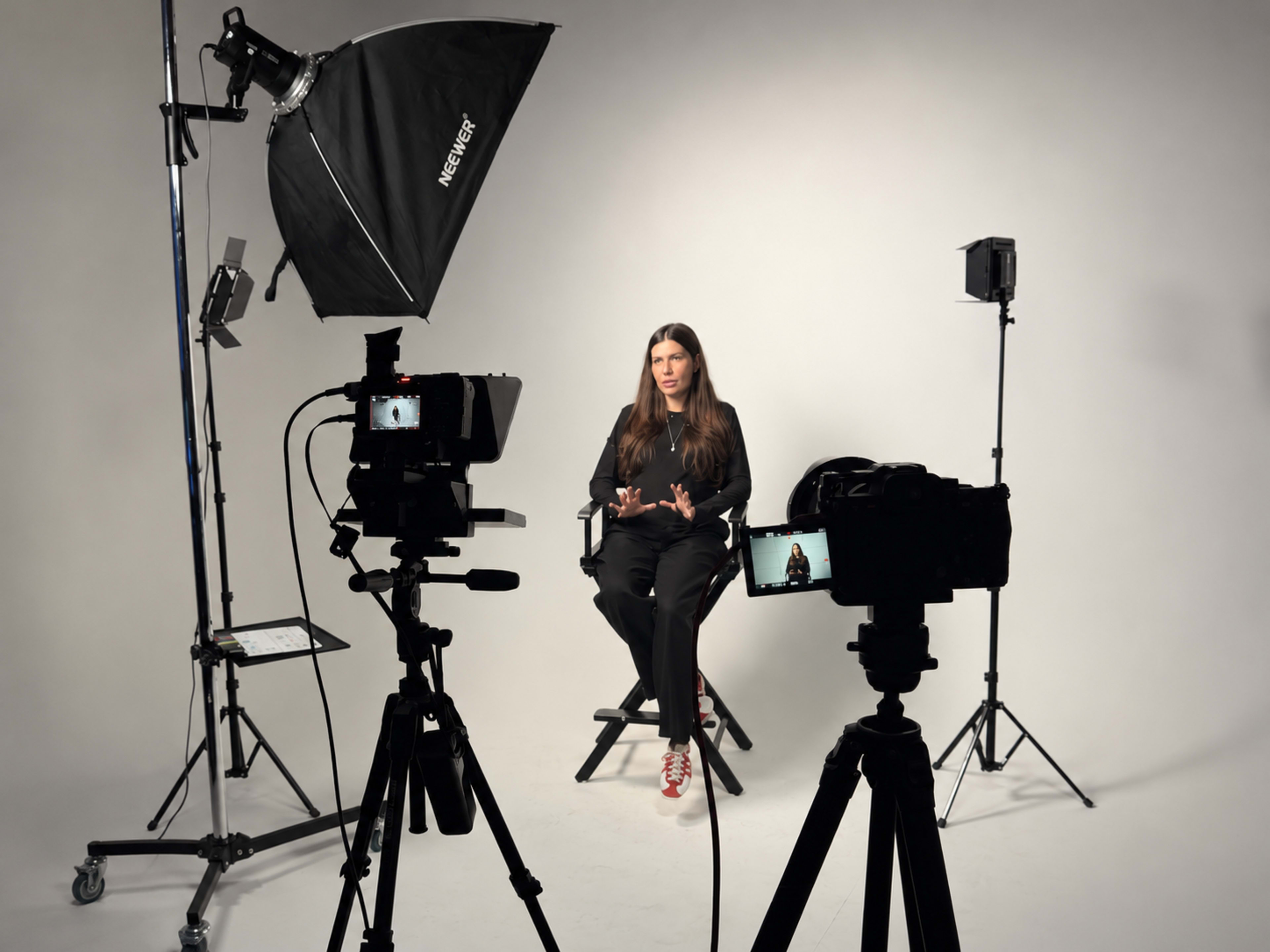 A woman sits on a director's chair in a photo studio, surrounded by multiple cameras and studio lights.