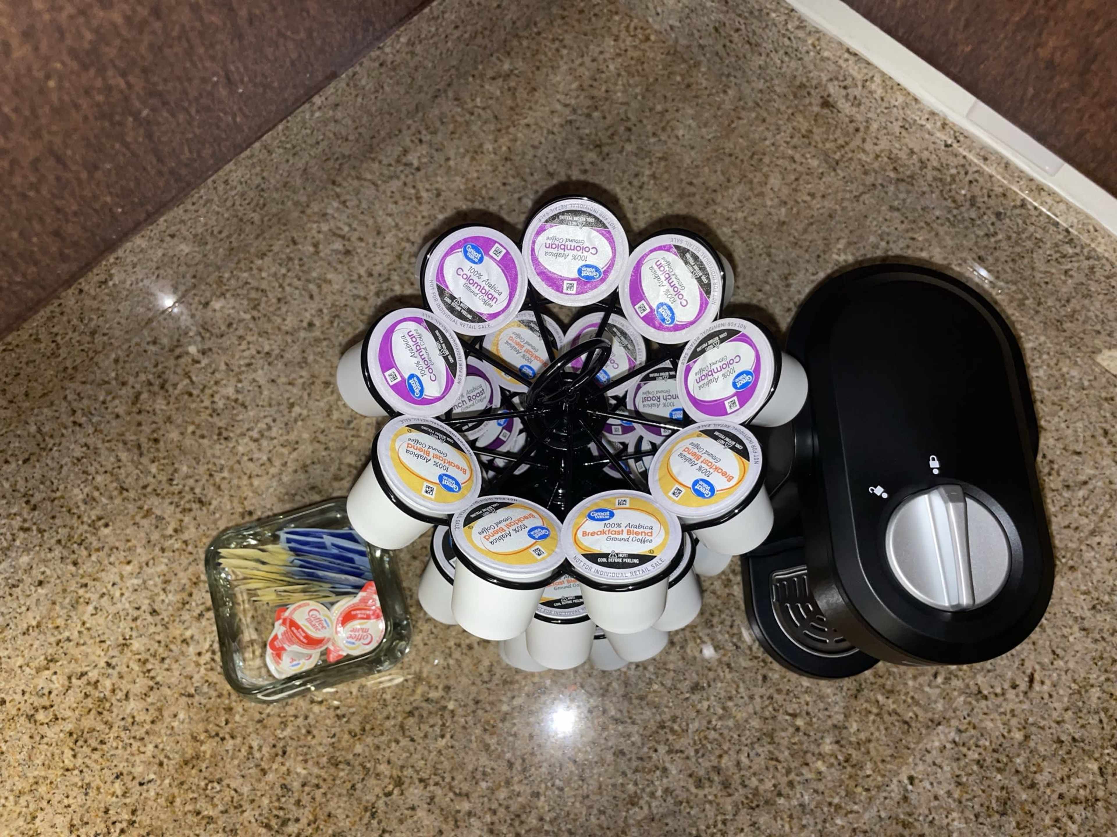 A coffee pod holder with multiple coffee pods arranged around a coffee maker, along with a small glass container holding utensils.